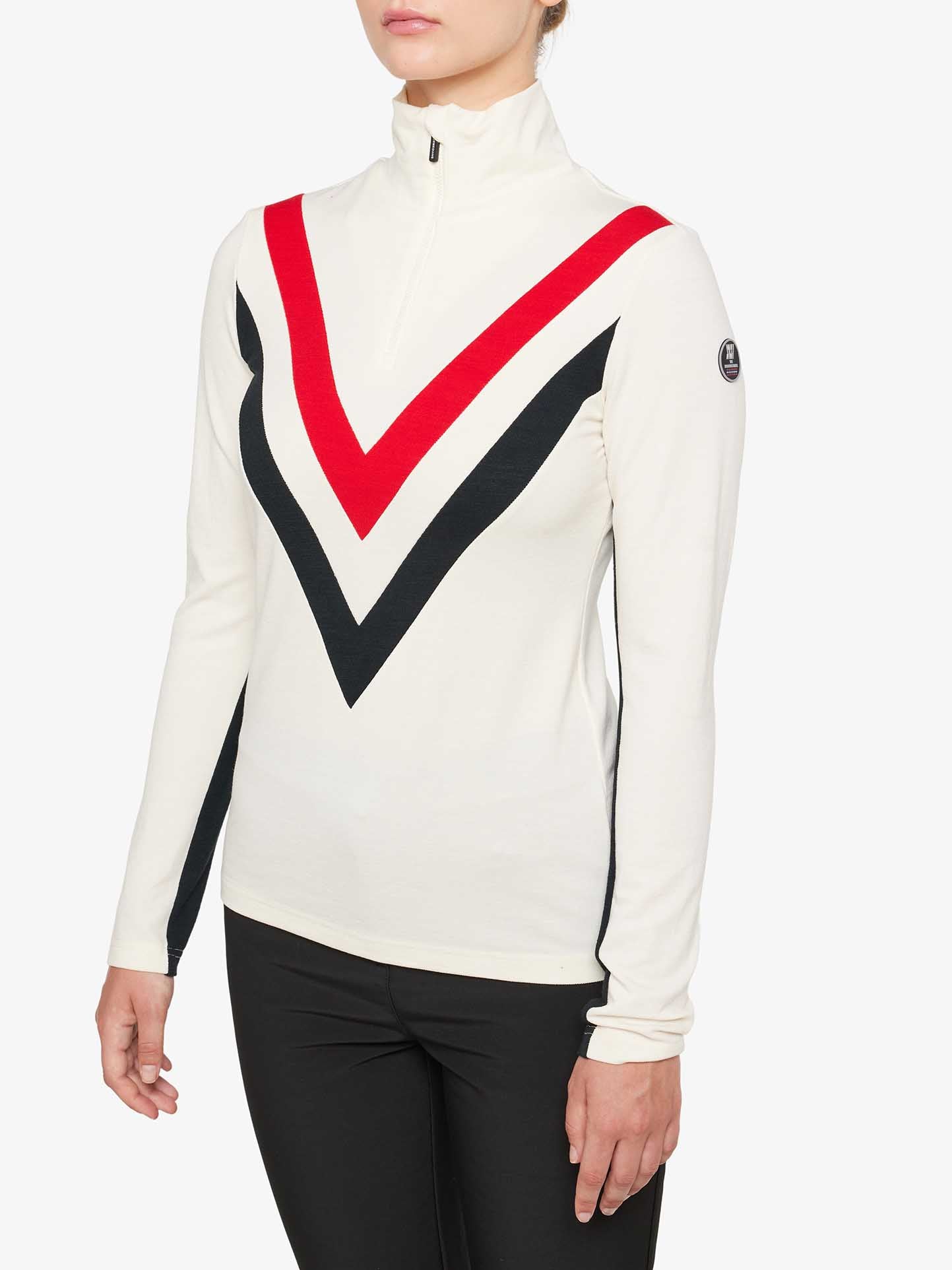 A person wearing a We Norwegians Voss Chevron ZipUp Women, featuring a white long-sleeve top with a high collar and half-zip front. The top boasts retro vibes with a bold V-shaped design in red and black on the chest and sleeves. Made from Super Fine Merino wool, it's paired stylishly with black pants. The We Norwegians logo is visible on the left sleeve.