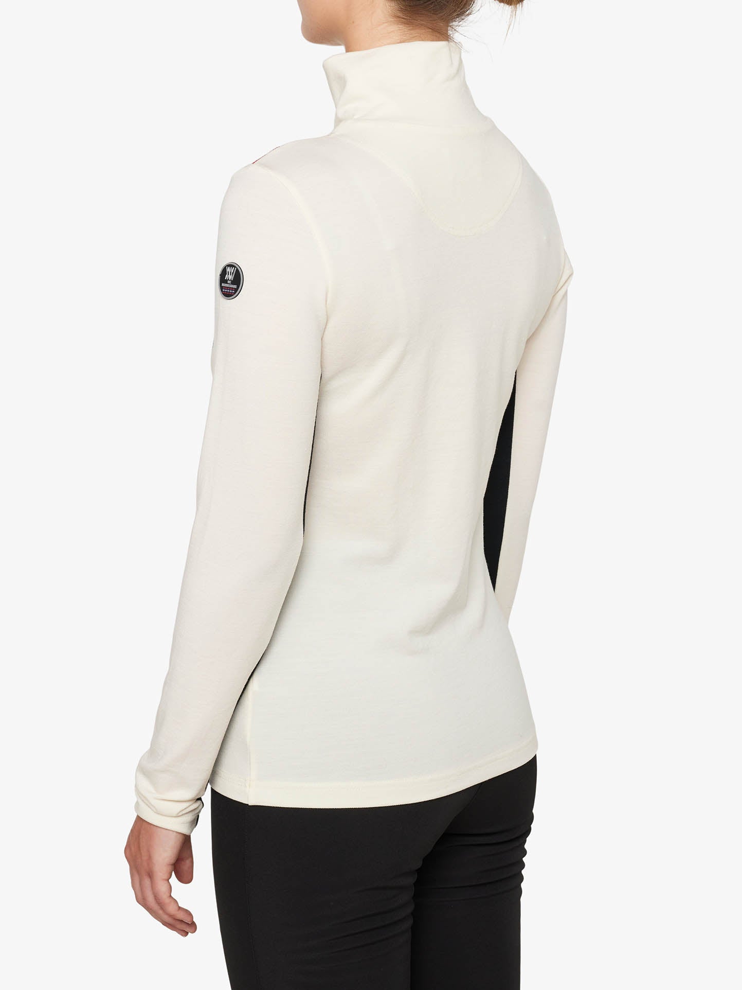 A person with light skin is wearing a white Voss Chevron ZipUp Women long-sleeve turtleneck by We Norwegians paired with black pants. The back view highlights the high-collared neckline and a small circular patch on the left sleeve, exuding retro vibes against the plain white background.