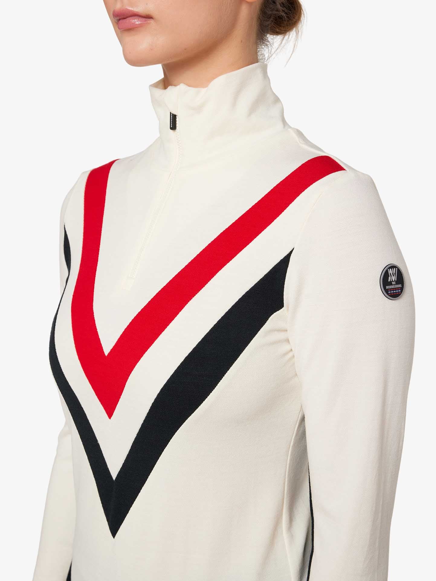 A close-up of a person wearing the We Norwegians Voss Chevron ZipUp Women, a long-sleeved, high-neck top with a half-zip closure. The top emits retro vibes with black and red chevron stripes pointing downwards across the chest and is crafted from Super Fine Merino wool. A circular patch with the logo adorns the upper left sleeve.