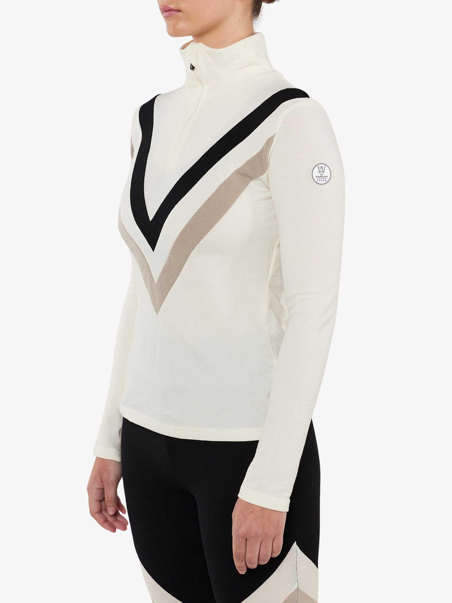 A person wearing the Voss Chevron ZipUp Women by We Norwegians, crafted from Super Fine Merino wool and showcasing black and beige V-shaped stripes on the front. The retro vibes of this white long-sleeve top with a high neck and partial zipper extend to the coordinating leggings that feature similar color accents. The background is plain white.