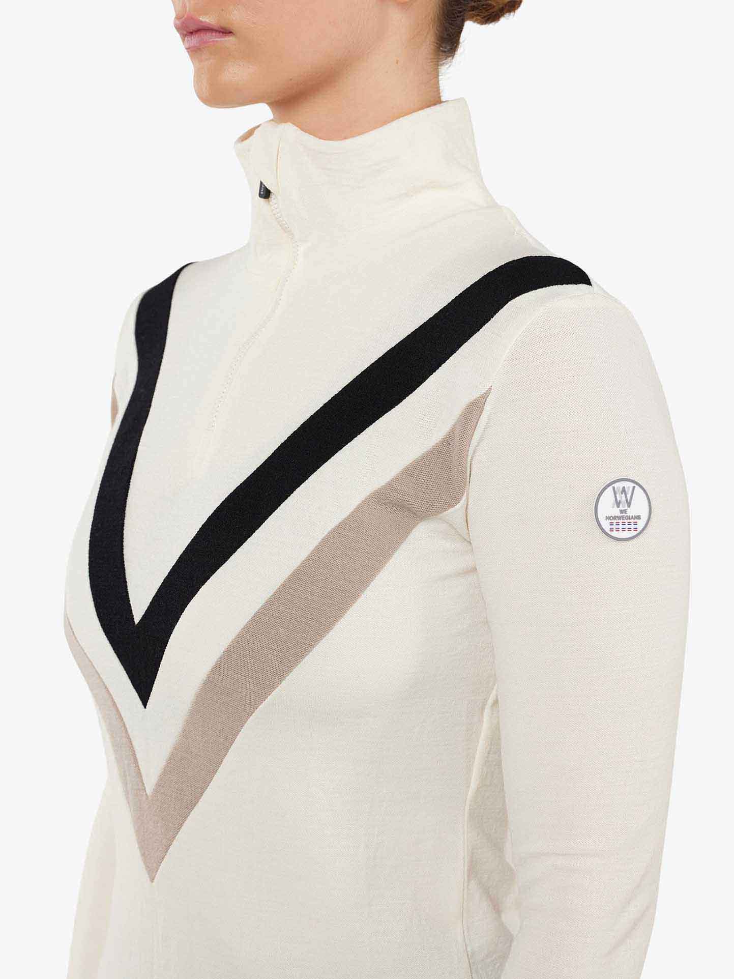 A person is wearing the Voss Chevron ZipUp Women by We Norwegians, which features a white design with a high collar and zip closure. The top showcases a striking large V-shaped pattern in black, beige, and gray on the chest. Constructed from super fine Merino wool, it also includes a small round logo on the sleeve, making it ideal for après ski elegance.