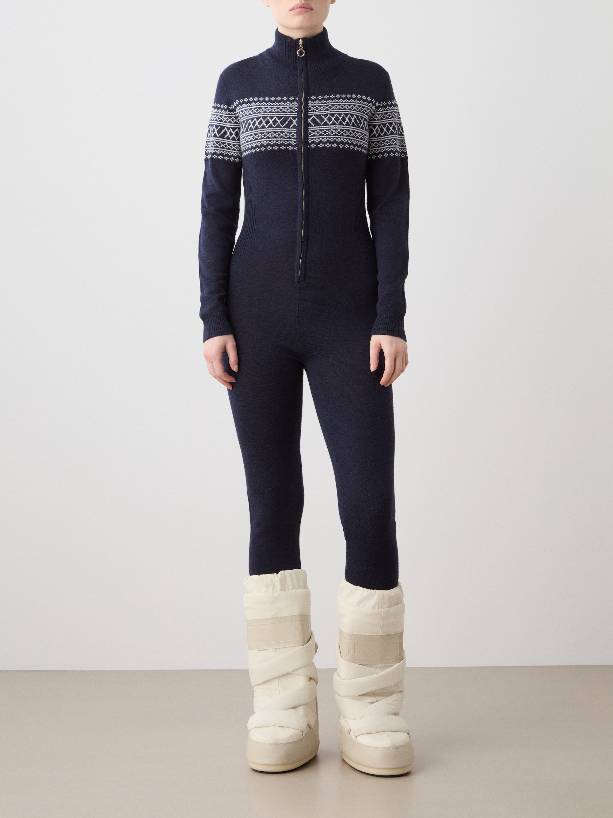 A person wears the We Norwegians Signature Jumpsuit Women, a navy blue, full-body knit made from washable merino with white Setesdal patterns on the chest and sleeves, paired with cream padded winter boots against a plain background.