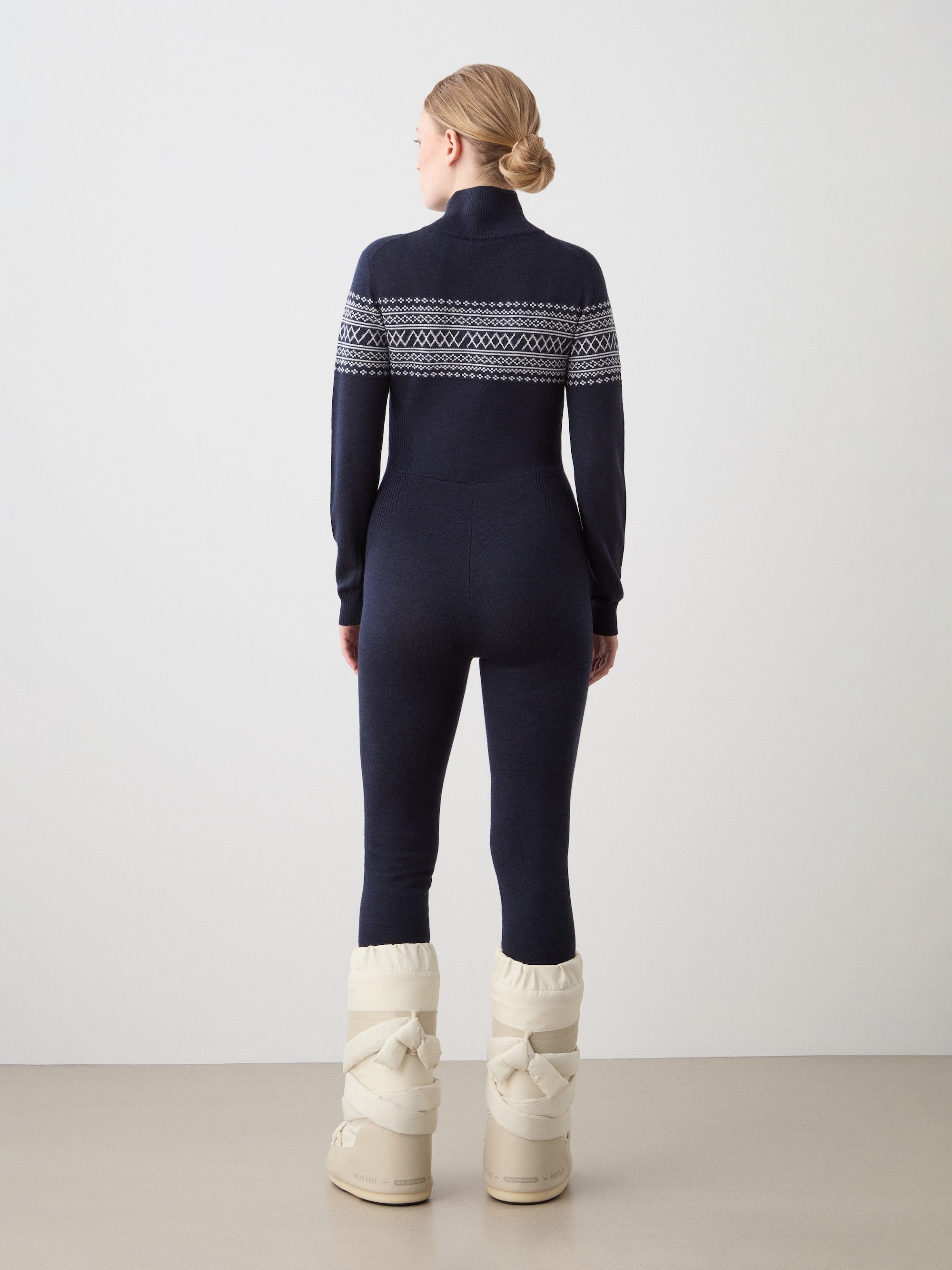 A person faces away, wearing the We Norwegians Signature Jumpsuit Women in navy with a Setesdal yoke, paired with cream winter boots, in a minimal indoor setting against a plain background.