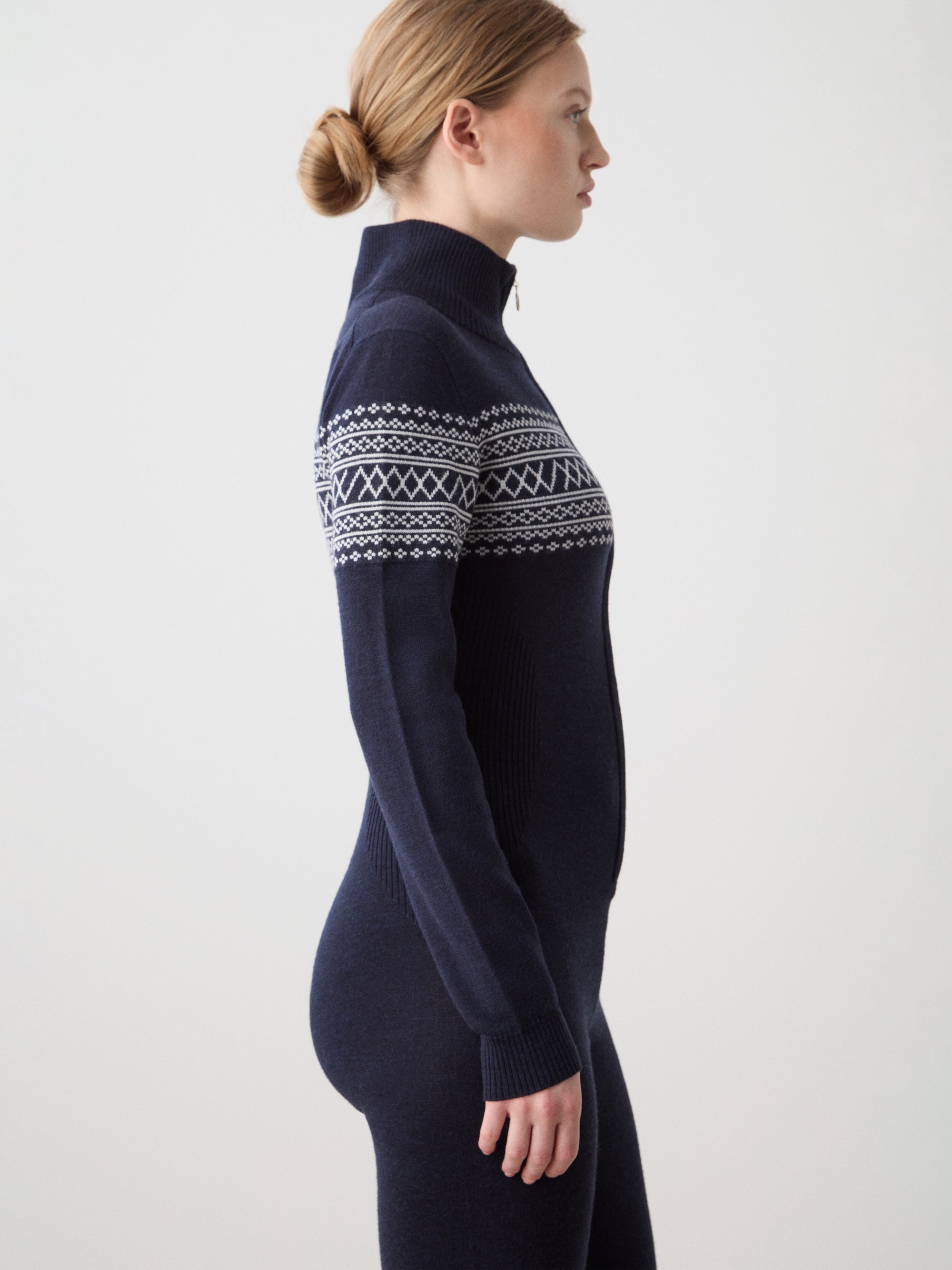 A woman with light hair tied back stands in profile against a plain light background, wearing the We Norwegians Signature Jumpsuit for women—a navy blue one-piece featuring a classic Setesdal pattern across the chest and arms.