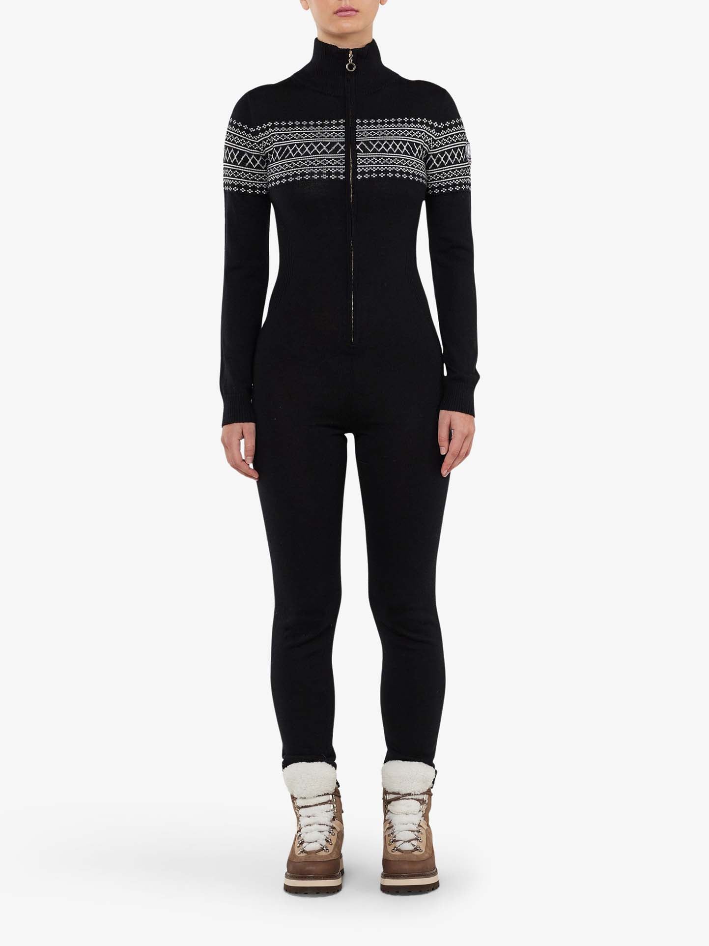 A person stands against a white background wearing the We Norwegians' Signature Jumpsuit Women, which features black, full-body design with Setesdal patterns on the upper chest, shoulders, and sleeves. The jumpsuit has a front zipper and is made from washable merino yarn. They are also wearing brown, fur-lined winter boots.