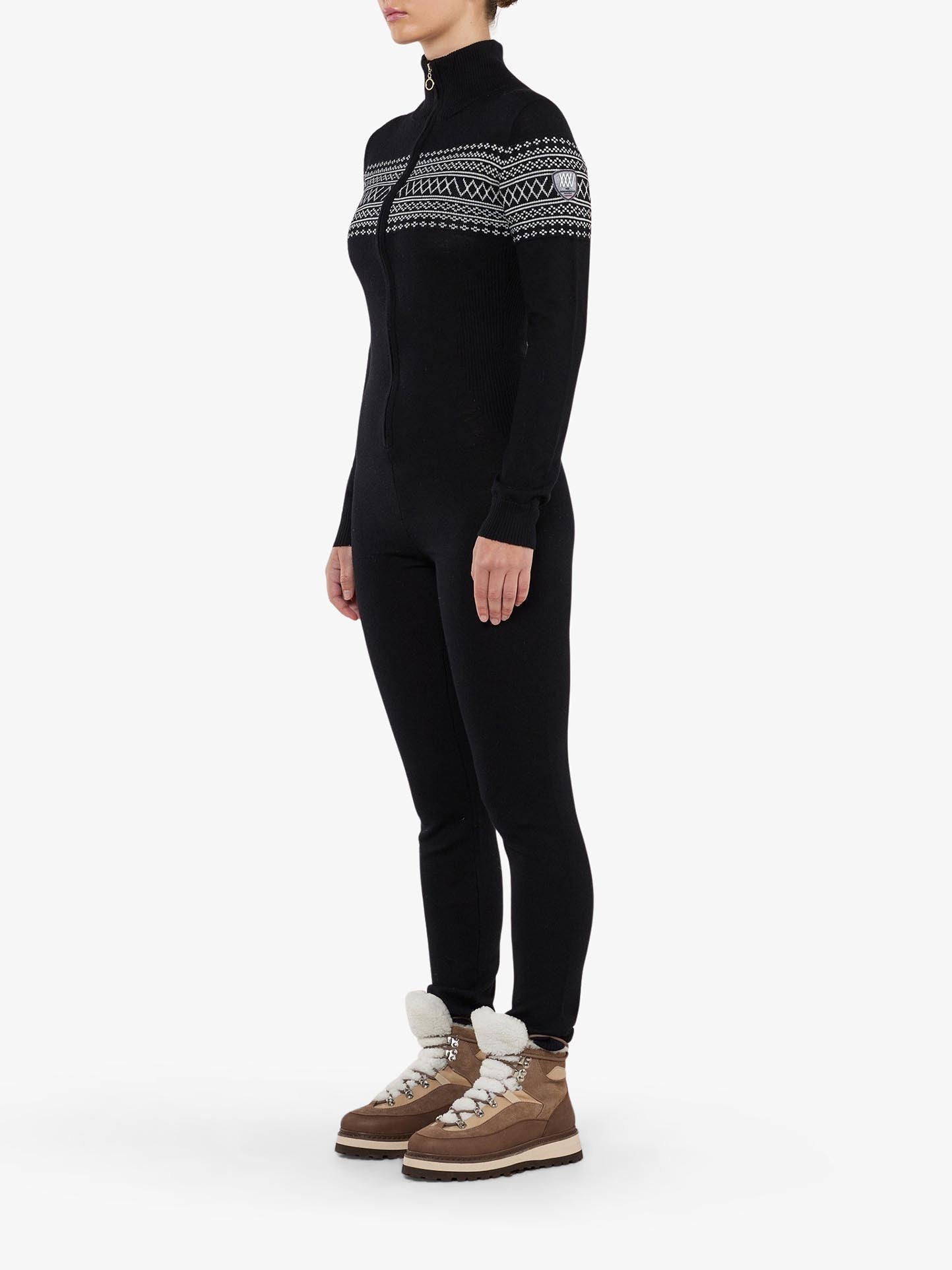 A person wearing the sleek black Signature Jumpsuit for Women by We Norwegians, featuring a full-body design with a zippered front and adorned with a white Setesdal pattern across the chest and shoulders. They are also sporting brown winter boots with white fur lining. The cozy ensemble stands out against a plain white background.