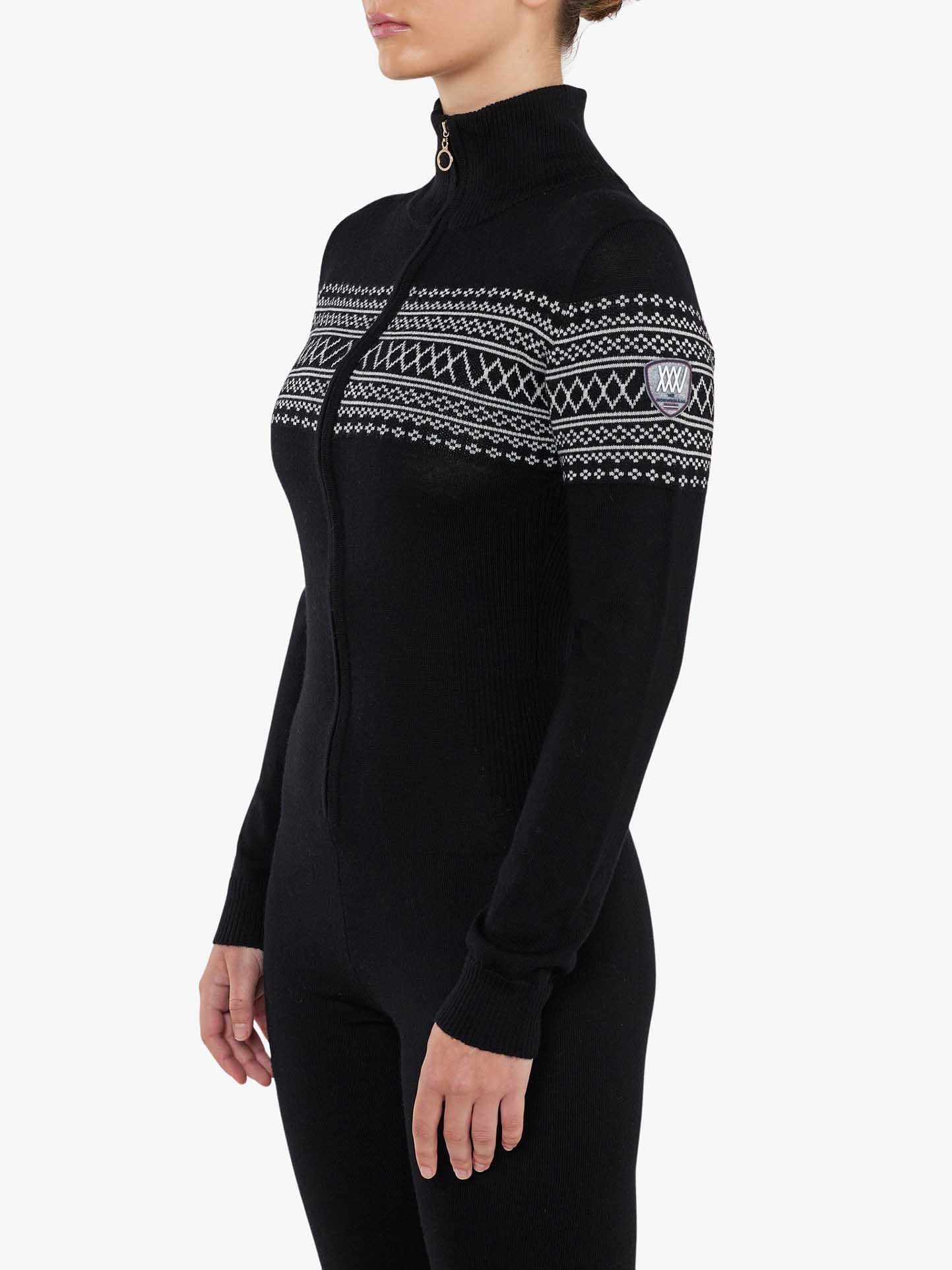 A person stands sideways wearing the We Norwegians' Signature Jumpsuit Women, a black, long-sleeved, zip-up bodysuit with a high collar. Crafted from washable merino yarn, this jumpsuit features white geometric patterns across the chest and upper arms.