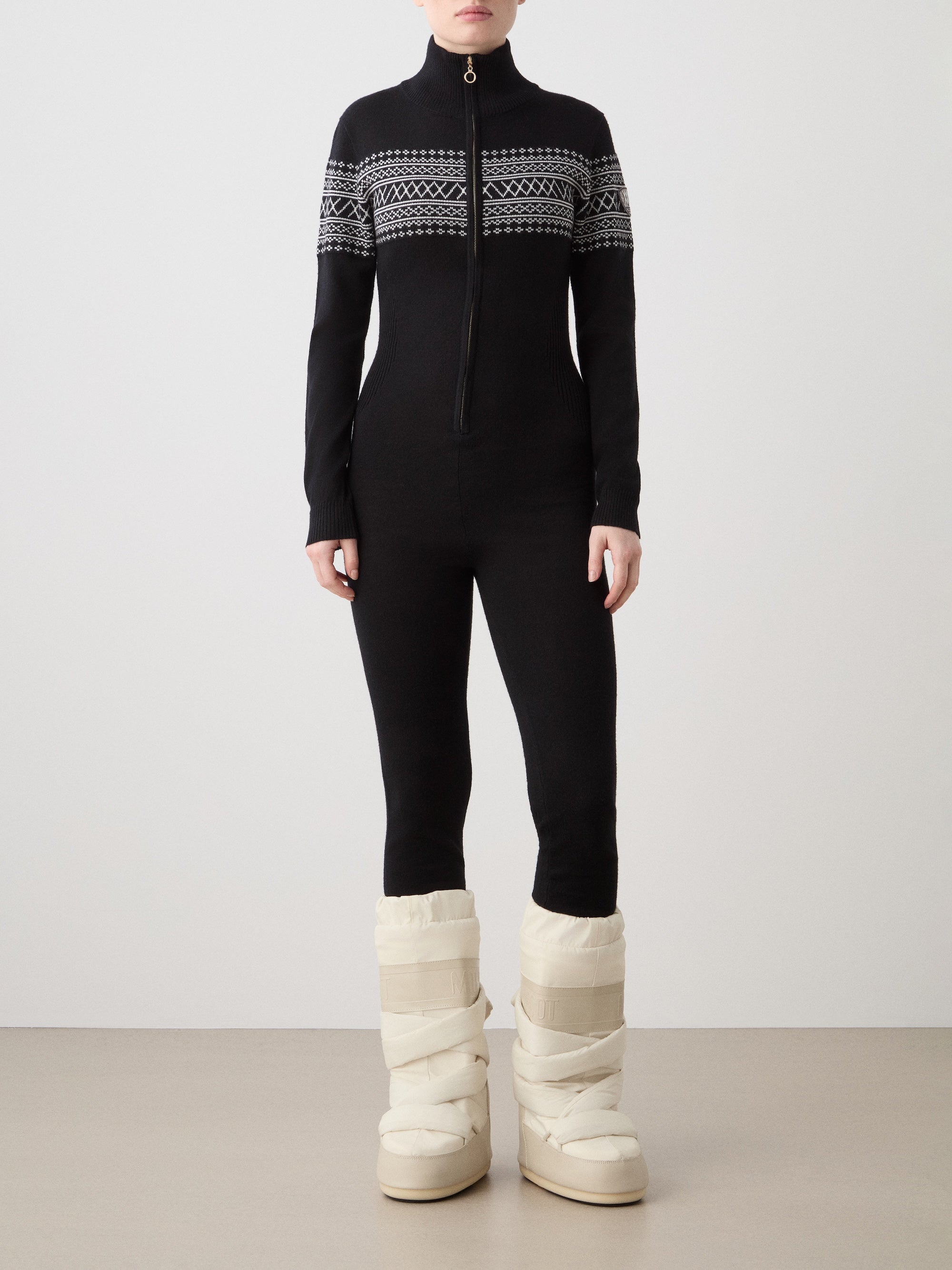 A woman models the We Norwegians Signature Jumpsuit—a fitted black one-piece with a white and gray geometric pattern—paired with tall puffy white winter boots, set against a plain light background.