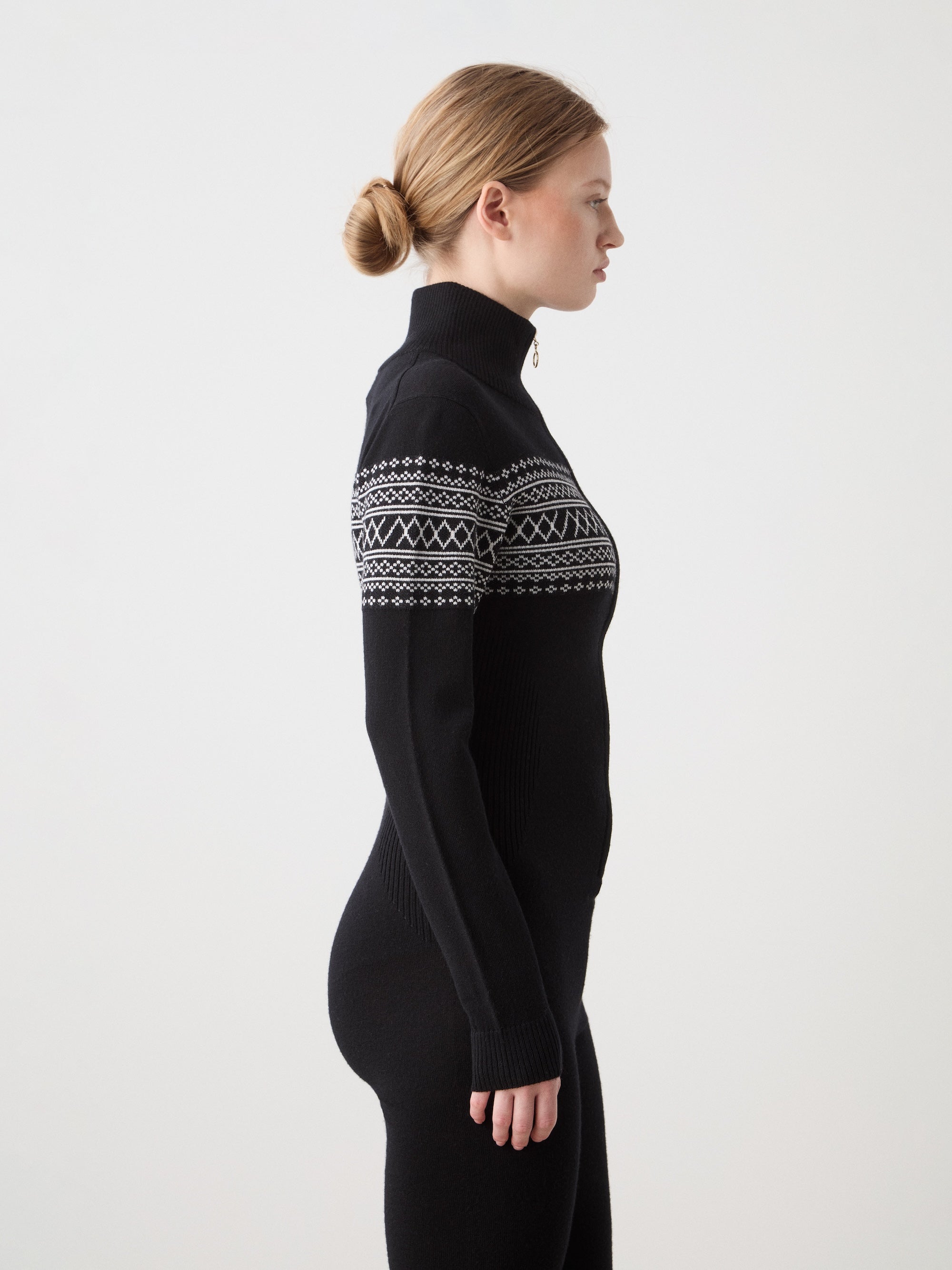 A woman with light skin and blonde hair in a bun stands in profile against a plain background, wearing the We Norwegians Signature Jumpsuit for women—a black après-ski piece with white geometric patterns, made from soft, washable merino yarn.