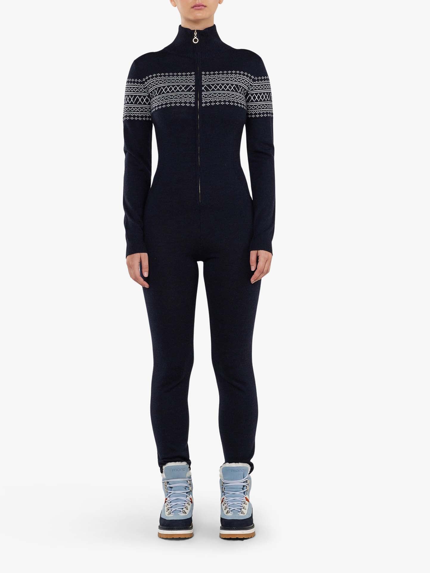 A person is wearing the Signature Jumpsuit Women by We Norwegians in navy blue, featuring a high neck and a front zipper. The upper section of the jumpsuit displays a white Setesdal pattern. They are also sporting light blue, lace-up winter boots, set against a plain white background.