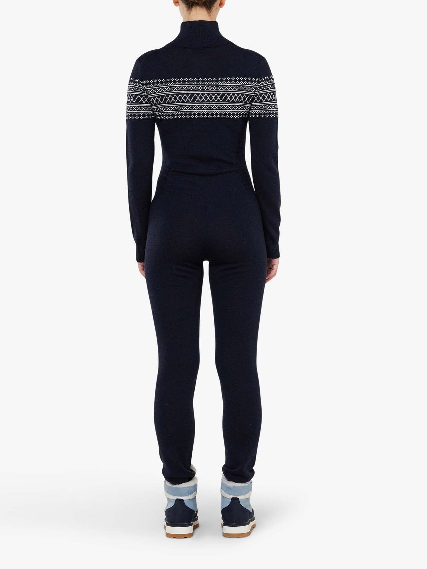 A person is standing with their back to the camera, wearing the Signature Jumpsuit Women by We Norwegians. This dark blue, long-sleeve jumpsuit showcases a modern take on the Setesdal pattern across the shoulders and upper back. Ideal as a base-layer ski outfit, it features light-colored rolled-up cuffs and matching dark blue shoes.