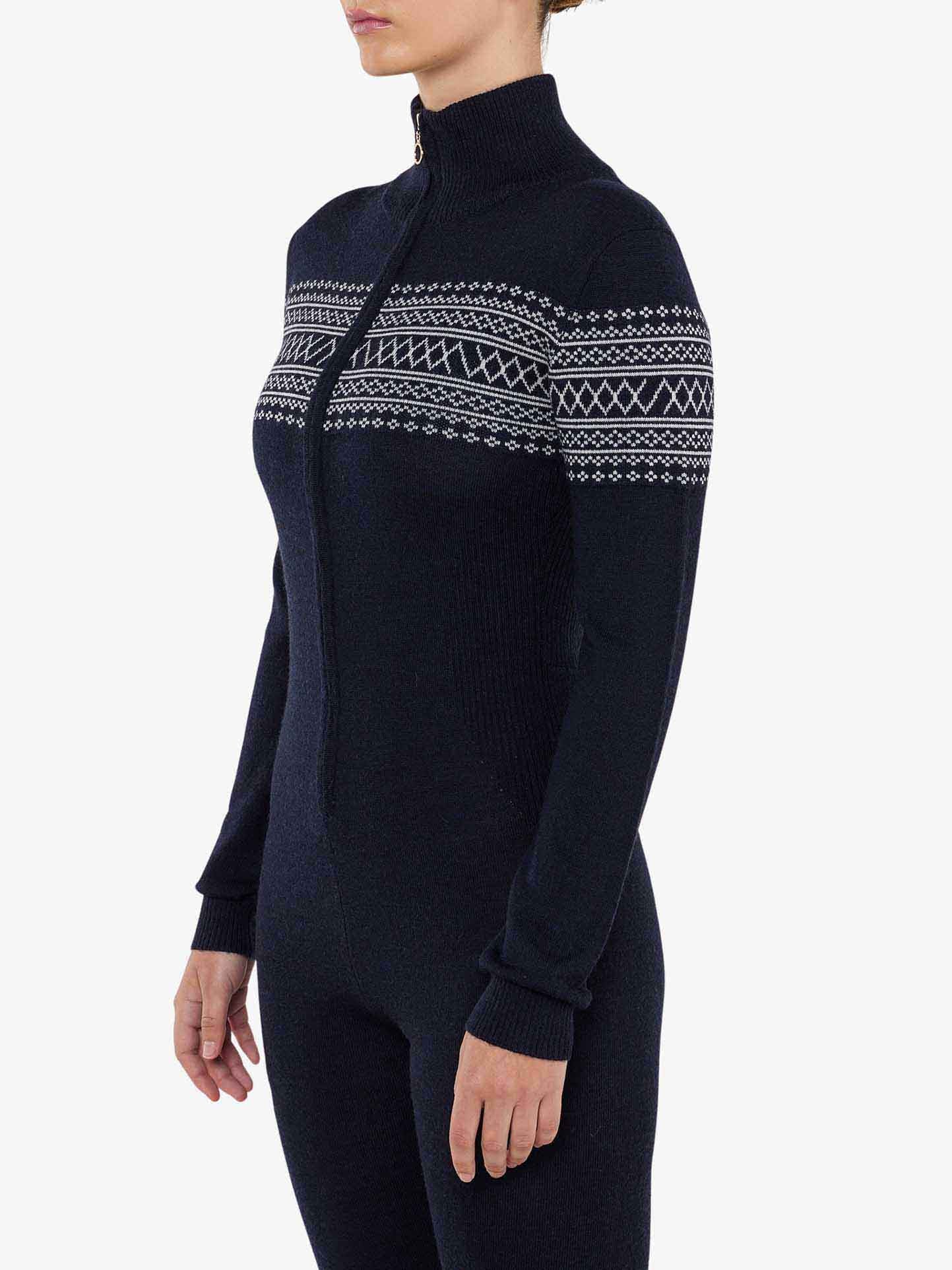 An individual is wearing the Signature Jumpsuit Women by We Norwegians, featuring intricate Setesdal patterns across the chest and sleeves. Crafted from washable merino yarn, this high-collared base-layer jumpsuit combines both style and practicality. The person is standing sideways with their hands relaxed by their side.