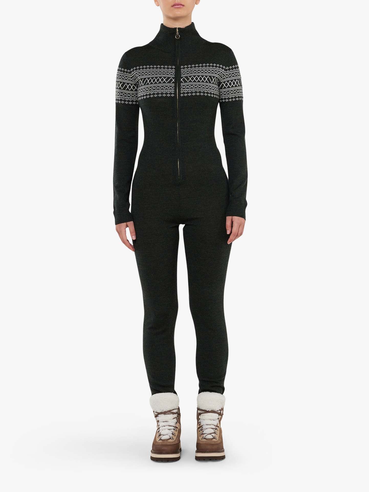 A person stands facing forward wearing the Signature Jumpsuit Women by We Norwegians, crafted from washable merino yarn. The black, long-sleeve jumpsuit features white geometric Setesdal patterns across the chest and sleeves. They are also wearing white socks and brown winter boots with fur lining. The background is white.