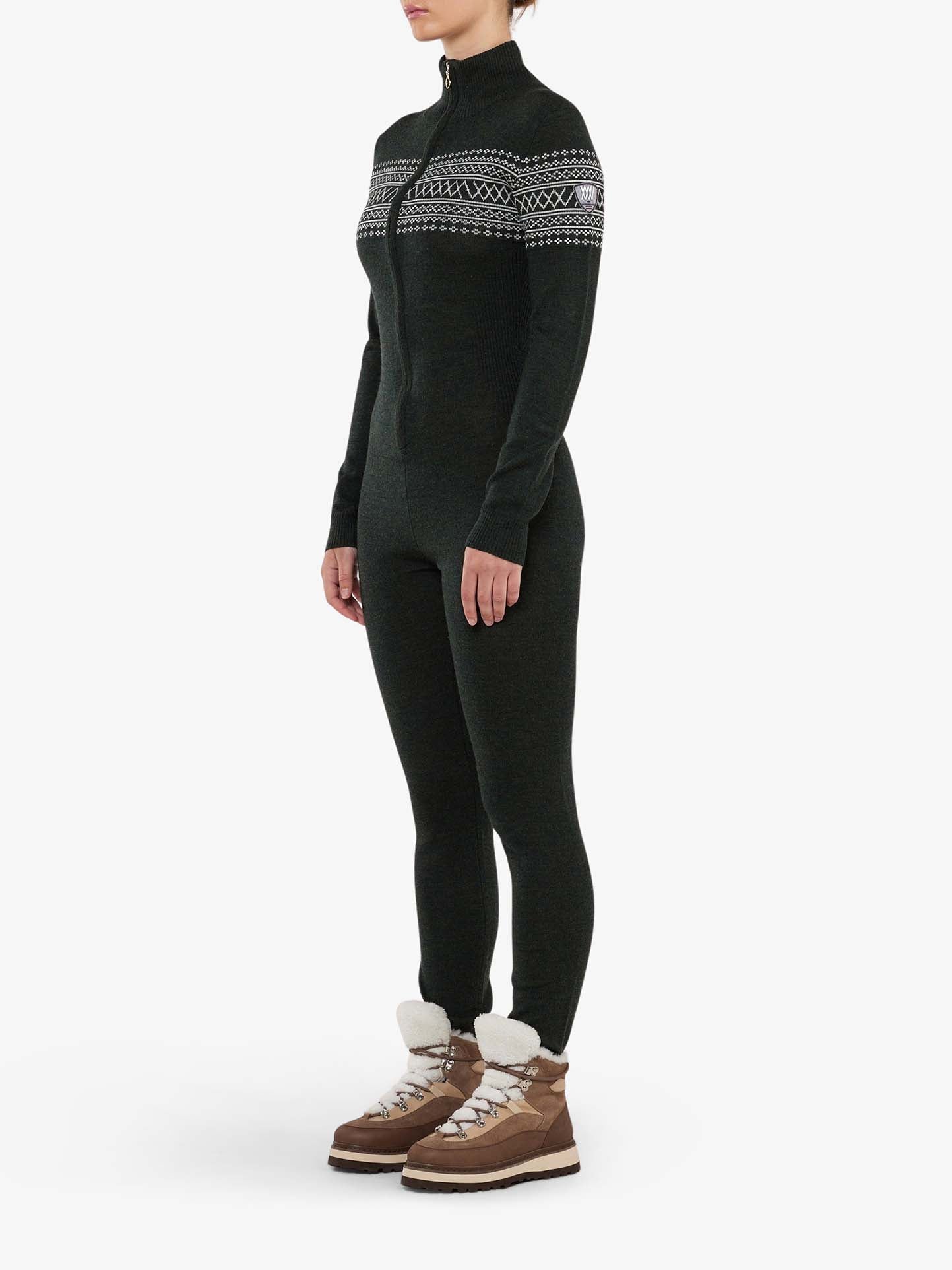 A person wearing the We Norwegians Signature Jumpsuit Women in dark green, featuring a full-length zip-up design with a white Setesdal pattern stripe across the chest and shoulders. Complementing their outfit are brown winter boots with white, fuzzy lining. The base-layer ski ensemble pairs perfectly against the plain white background.