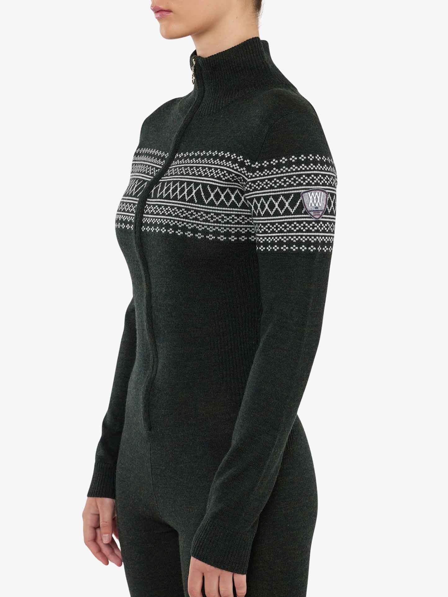 A person with short hair is shown from the side, wearing a fitted, dark-colored Signature Jumpsuit for women by We Norwegians. This stylish jumpsuit features a long-sleeved, zip-up turtleneck design adorned with a white Setesdal pattern across the chest and upper sleeves. The background is plain and light-colored.