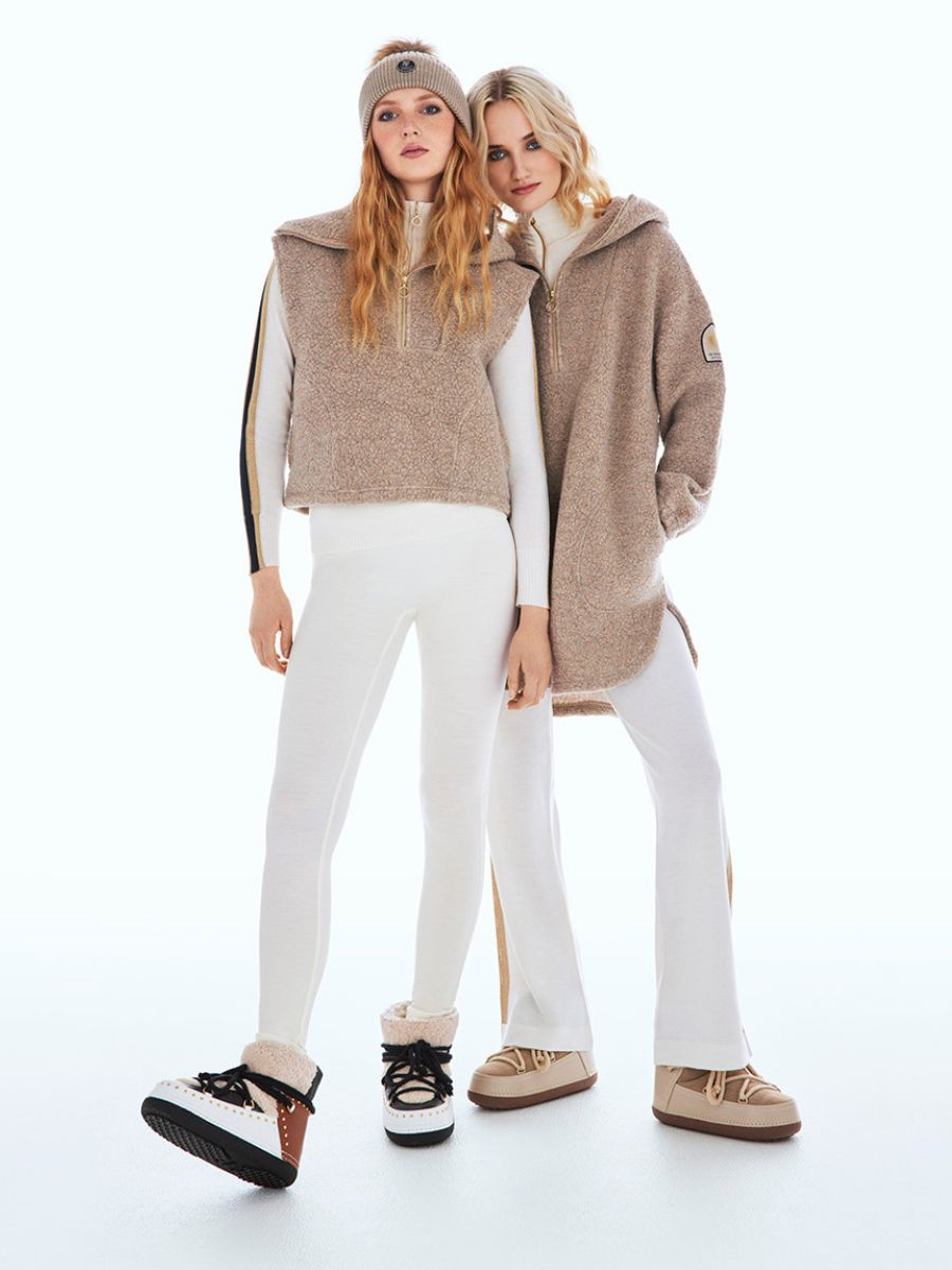 Two women are standing side by side against a white background, dressed in cozy, winter-inspired outfits from the We Norwegians Alta collection. One wears the Alta Shearling Hoodie Women while the other dons a sleeveless vest with a beanie. Both pairs of boots match their outfits perfectly.