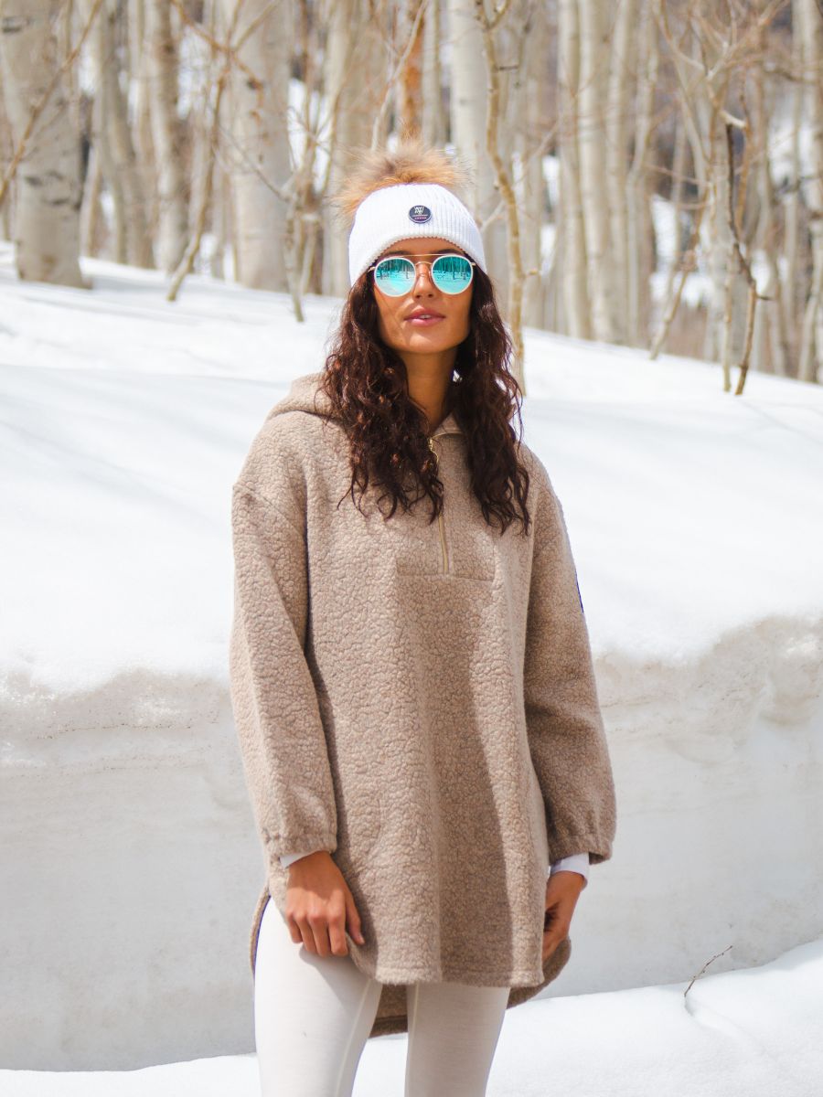 A person stands in a snowy forest, wearing an "Alta Shearling Hoodie Women" by We Norwegians, white leggings, a white headband with a pom-pom, and reflective sunglasses. The trees are bare, and the ground is covered in deep snow, creating a serene winter scene.