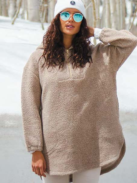 A person stands outdoors in a snowy landscape wearing a beige, cozy-looking oversized Alta Shearling Hoodie Women from We Norwegians, a white beanie, and reflective blue sunglasses. They have one hand to the back of their head and the other relaxed by their side. Trees are visible in the background.