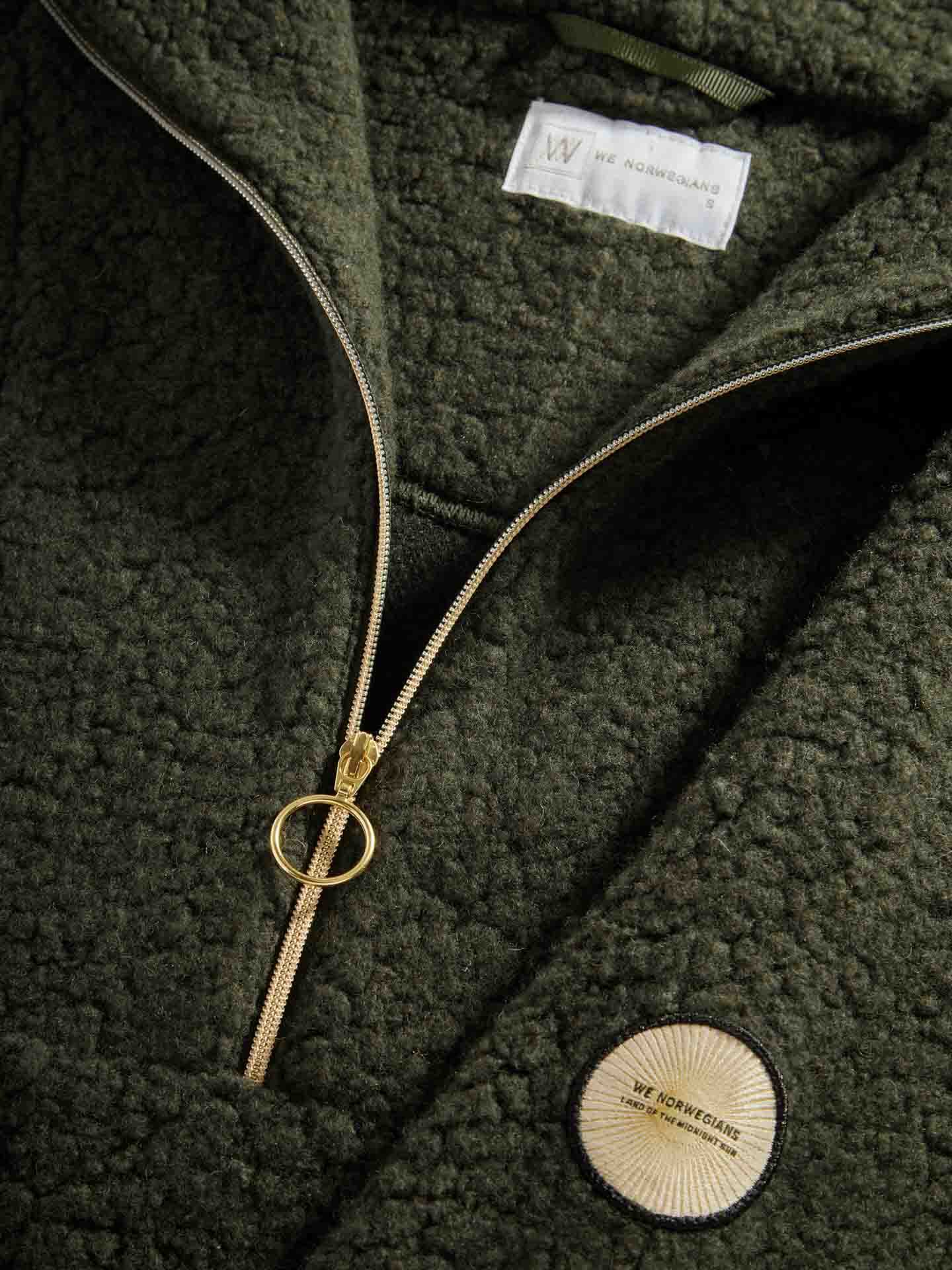 Close-up of a green, textured fabric Alta Shearling Hoodie Women by We Norwegians, featuring a gold zipper partially unzipped with a circular zipper pull and a label underneath the collar. A round patch with white text on a beige background is sewn near the zipper.