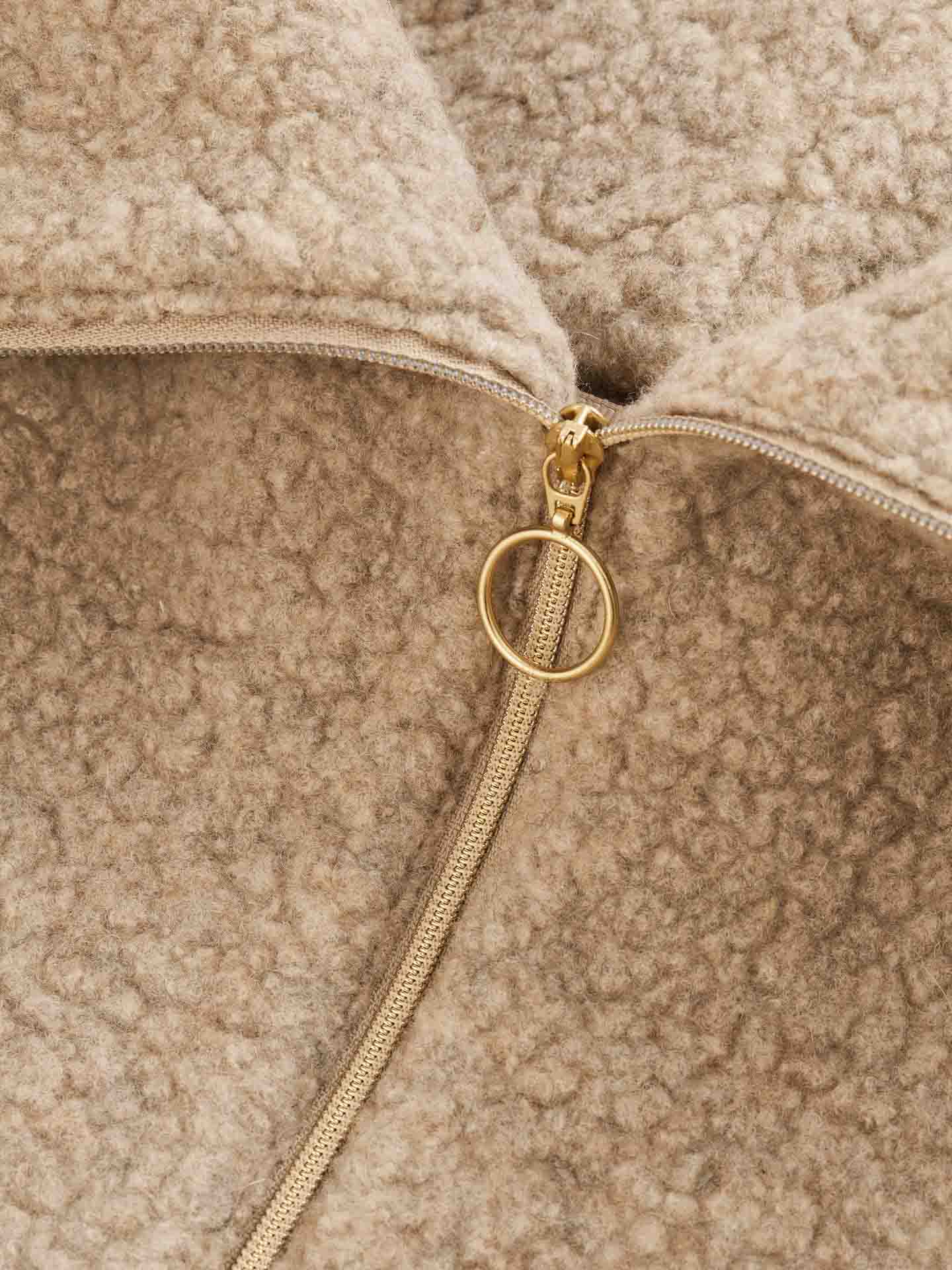 Close-up of the Alta Shearling Hoodie Women by We Norwegians, showcasing its tan-colored teddy fleece fabric with a partially open metal zipper featuring a circular pull tab. The texture of the fabric appears soft and plush, reminiscent of the cozy textures found in après-ski wear.