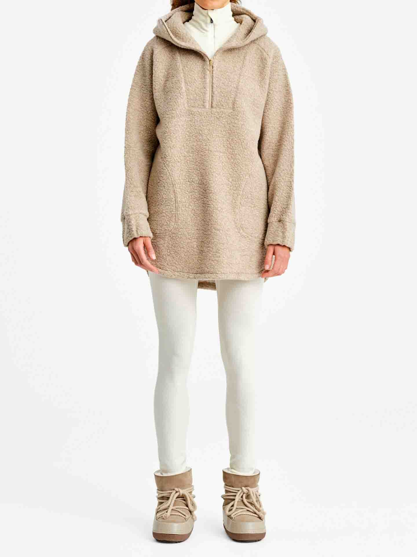 A person is wearing the Alta Shearling Hoodie Women by We Norwegians, featuring a beige sherpa fleece with a hood, half-zip front, and kangaroo pocket, making it perfect for après-ski wear. They are also dressed in white leggings and beige ankle boots against a plain white background.