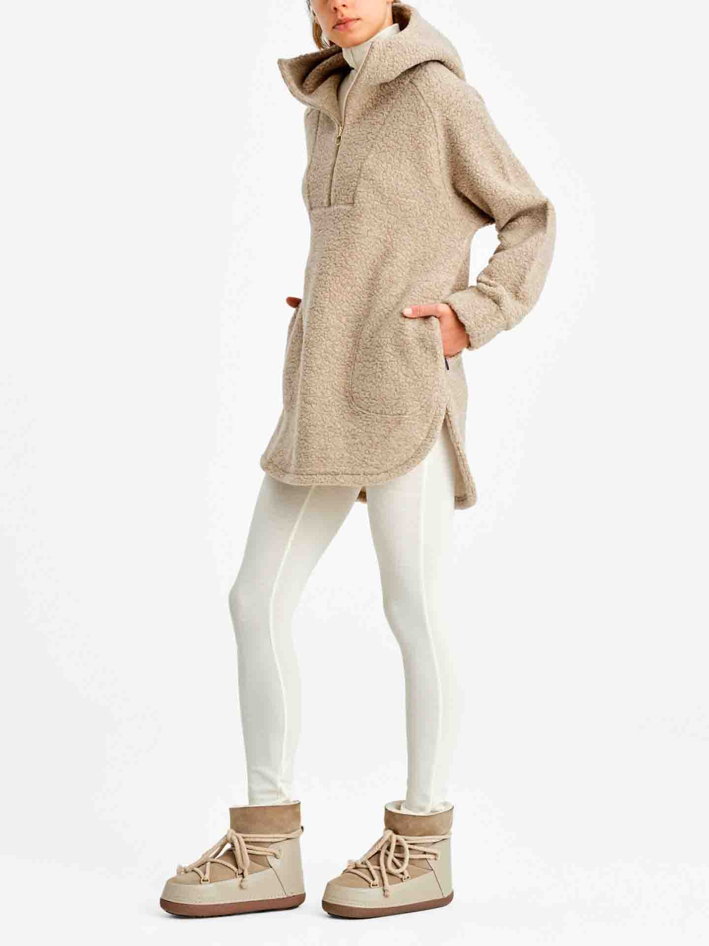 A person wearing the We Norwegians Alta Shearling Hoodie for Women, featuring an over-sized beige design with a zippered neckline and front pockets, paired with white leggings and beige, ankle-high, lace-up boots. The background is plain white.