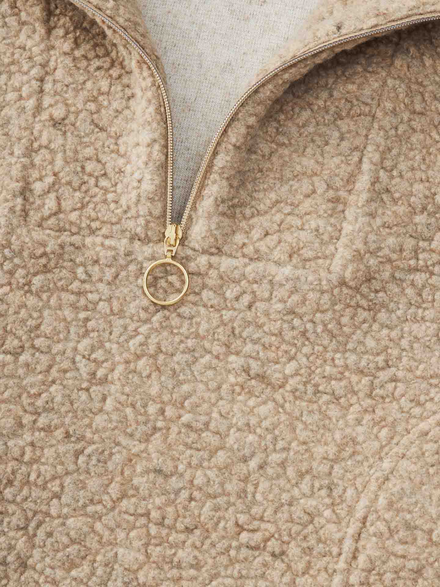 Close-up of a beige, textured fleece Alta Hooded Vest Women from We Norwegians, featuring a partial zip with a gold ring pull. The fabric has a soft, Merino wool-like appearance, and the zipper is not fully closed, revealing a light-colored layer underneath.