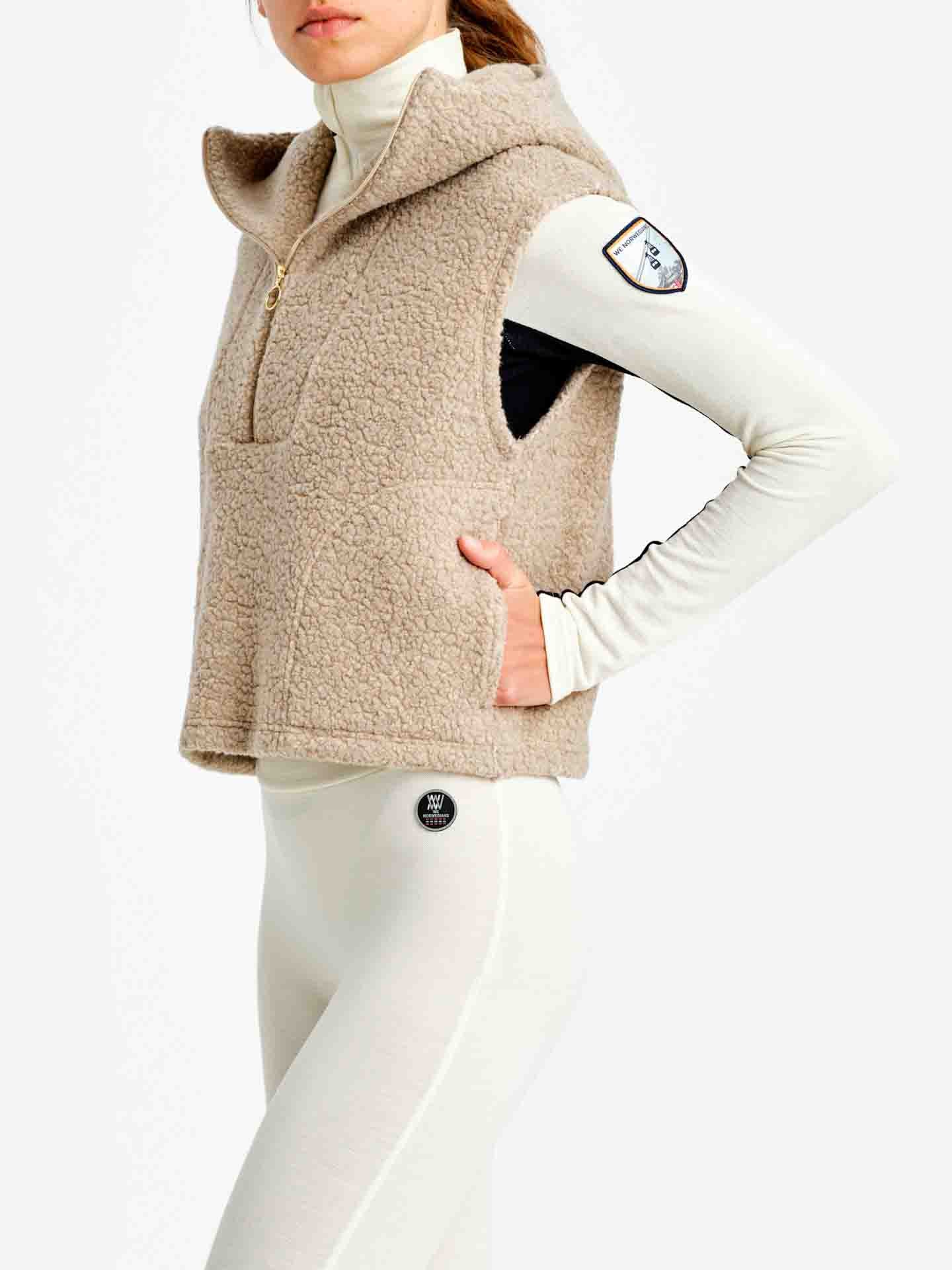 A person wearing a light beige Alta Hooded Vest Women from We Norwegians, featuring a zipper front, over a white turtleneck and white leggings. The vest boasts a high collar, two pockets, and an emblem patch on the left arm. Crafted from Merino wool, it merges comfort and style effortlessly against the plain white background.