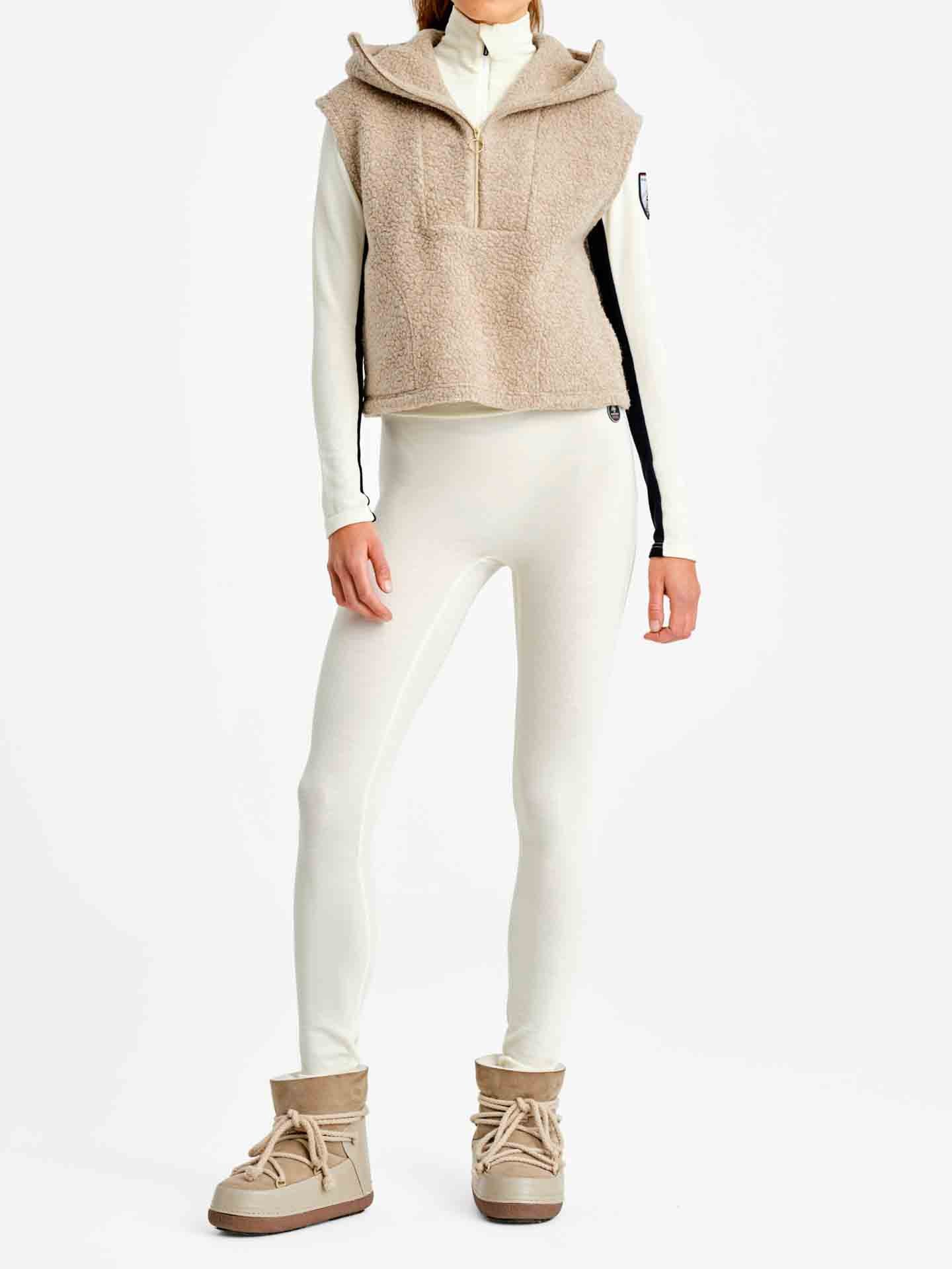 A person is standing against a plain white background wearing a cream-colored, full-body, form-fitting outfit. Over this, they have the "Alta Hooded Vest Women" from We Norwegians. They are also wearing beige, ankle-high boots with round toes and lace-up details.