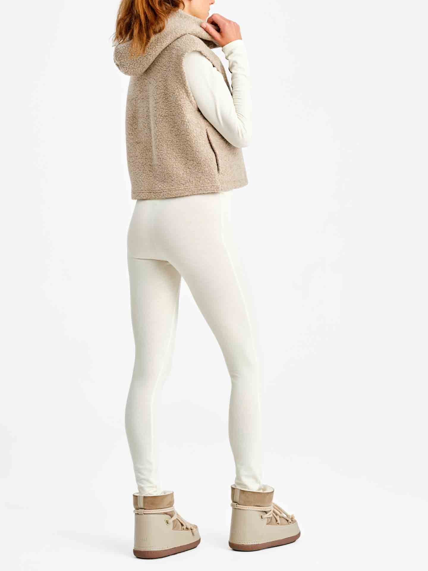 A person stands in a light-colored outfit featuring the We Norwegians Alta Hooded Vest Women, a sleeveless beige fleece vest, layered over a long-sleeve white top and fitted white pants. They are also wearing beige snow boots and have one hand touching their lips. The backdrop is a plain white background.