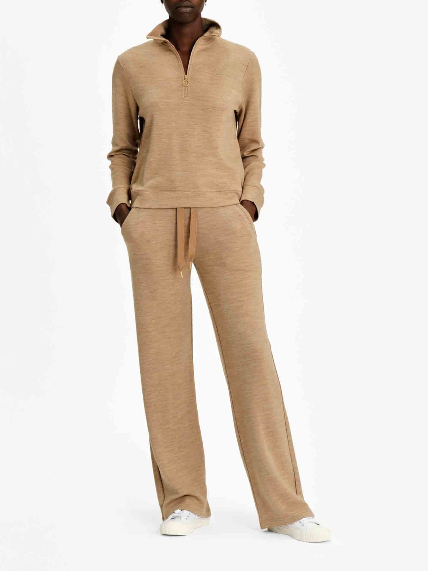 A person is wearing the Limited Time Bundle: Tind ZipUp & Tind Straight Leg Pant Women from We Norwegians in a beige color, made of luxurious merino wool. They have their hands in the pants' pockets and are standing against a plain, white background. The outfit is paired with white sneakers.