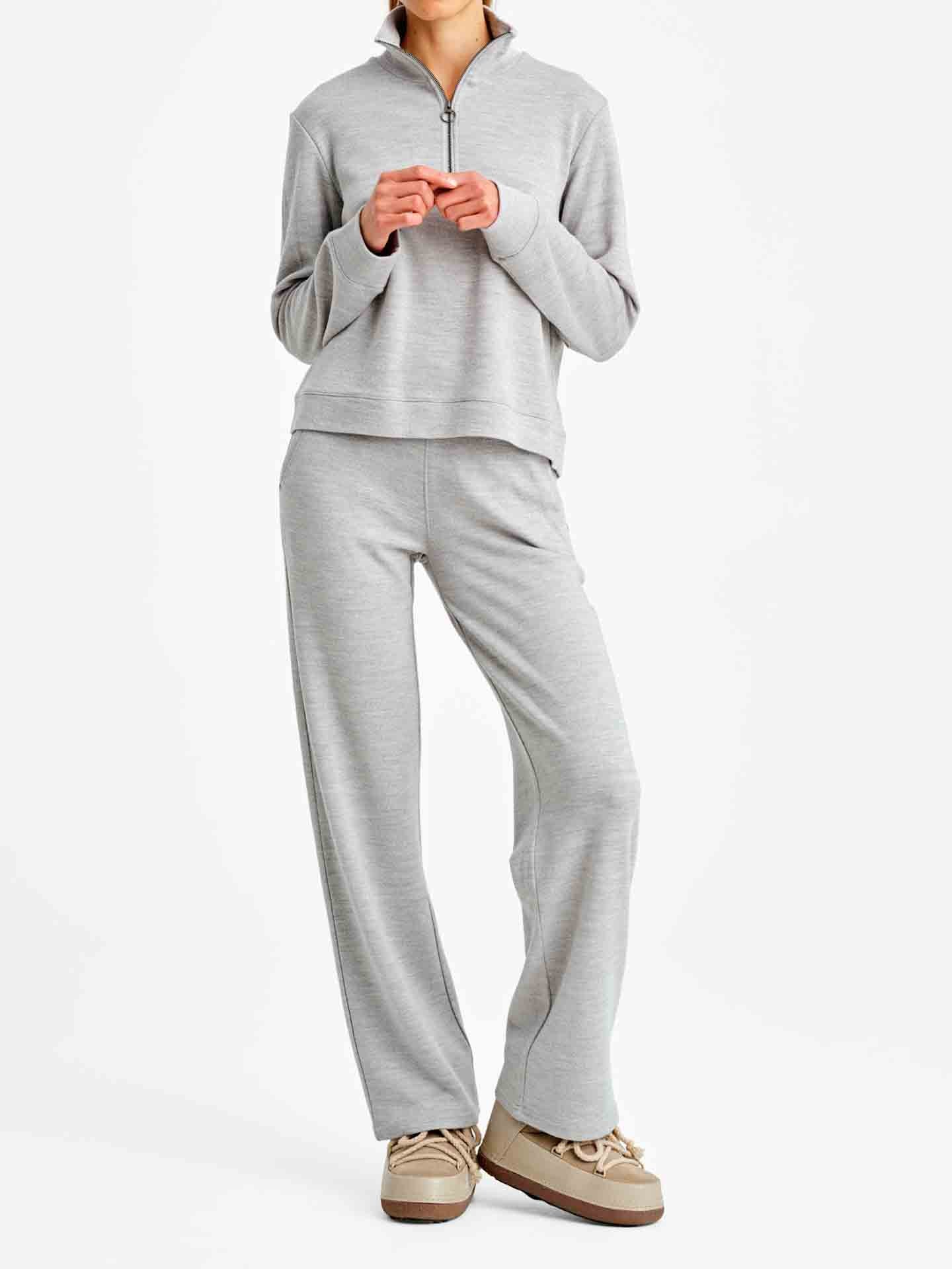 A person is standing against a plain white background wearing the Tind ZipUp Sweater in light gray and matching Tind Straight Leg Pants from the Limited Time Bundle by We Norwegians. They are also wearing beige shoes with a white sole. Their hands are clasped together at chest level.