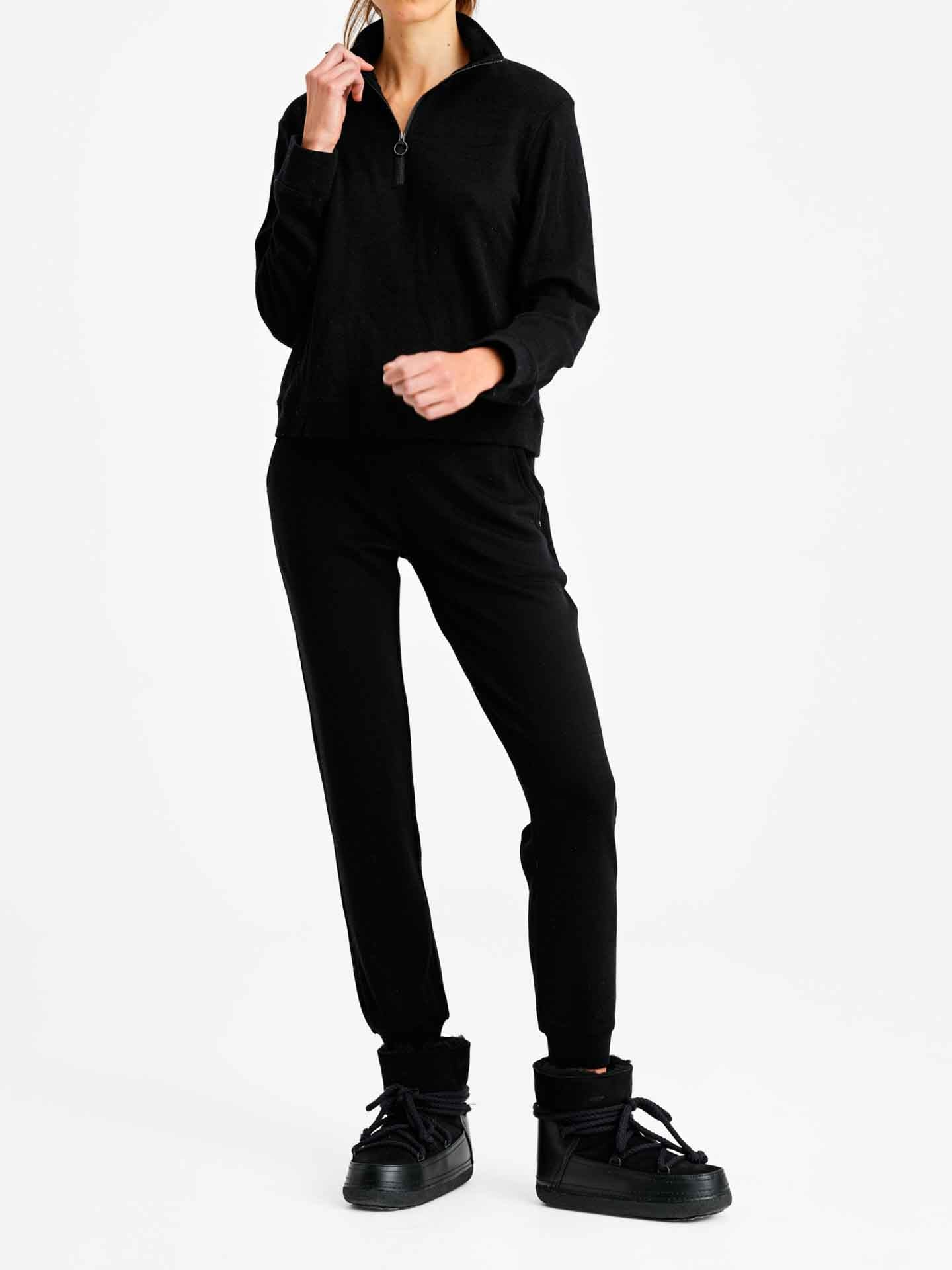 A person dressed in the all-black "Limited Time Bundle: Tind Zip Up & Tind Jogger Women" ensemble from We Norwegians, posing against a plain white background. They are also wearing black high-top sneakers with thick soles and holding something up near their face.