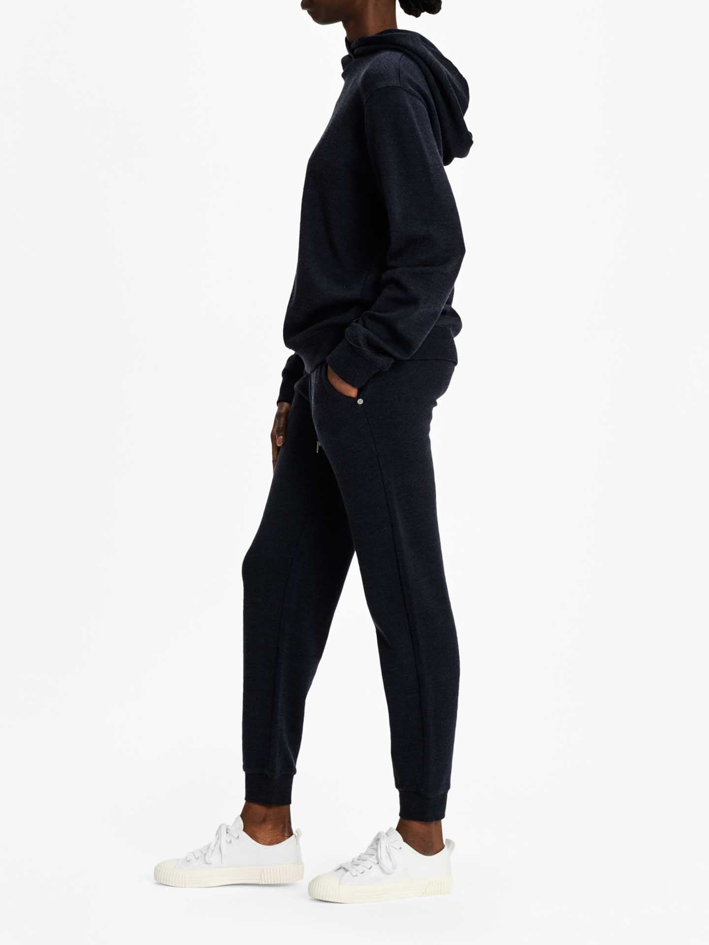 A person is shown standing in profile against a plain white background. They are wearing a dark-colored hoodie and matching Tind Jogger Women Navy pants by We Norwegians, with hands in pockets. The outfit is paired with white sneakers, showcasing a relaxed elegant style. The individual has short hair styled in a bun.