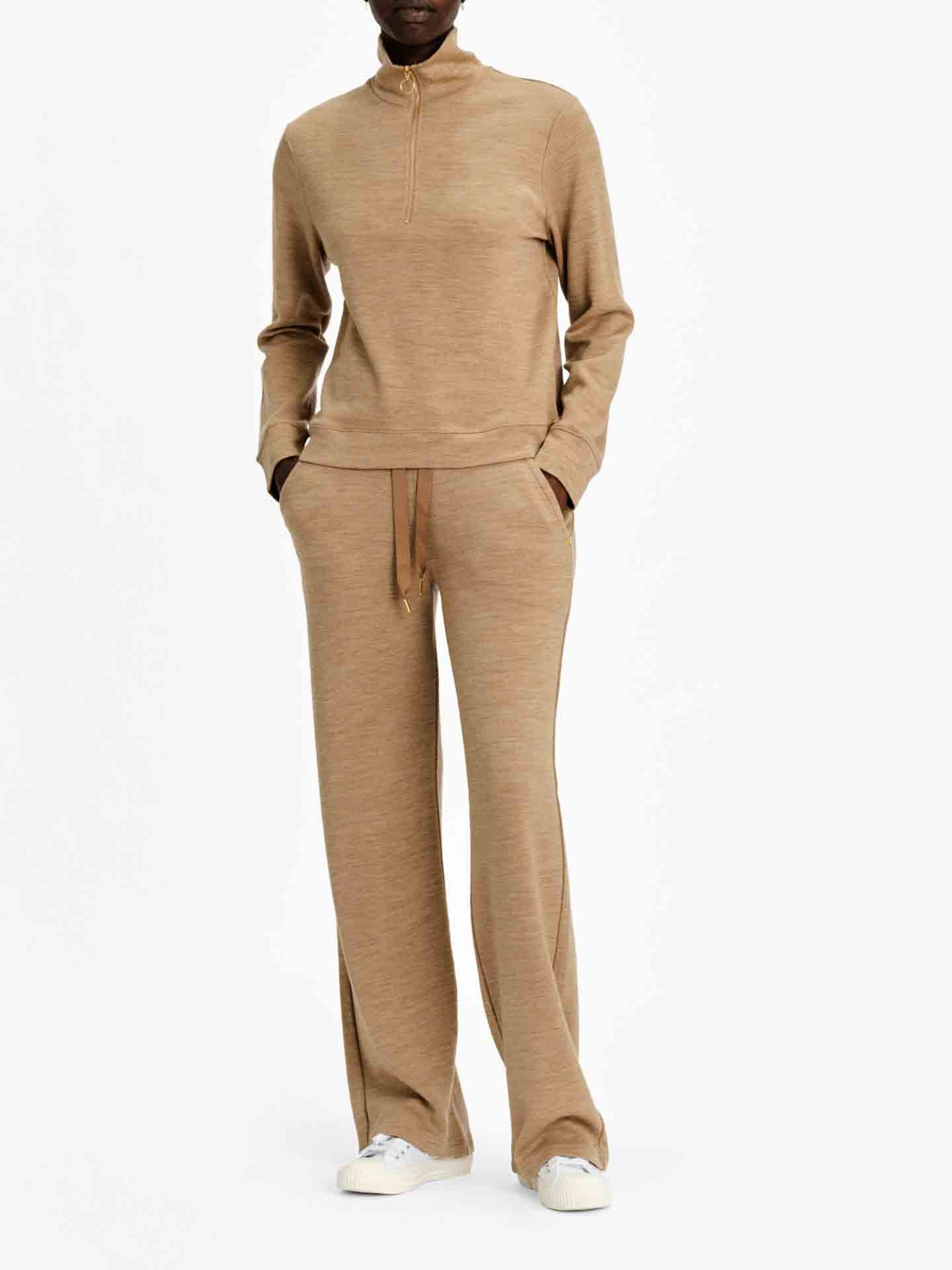 A person is wearing a beige zip-up long-sleeve top crafted from jacquard knit, paired with We Norwegians' Tind Straight Leg Pants in camel. They have their hands in their pockets and are wearing white sneakers against a plain white background.