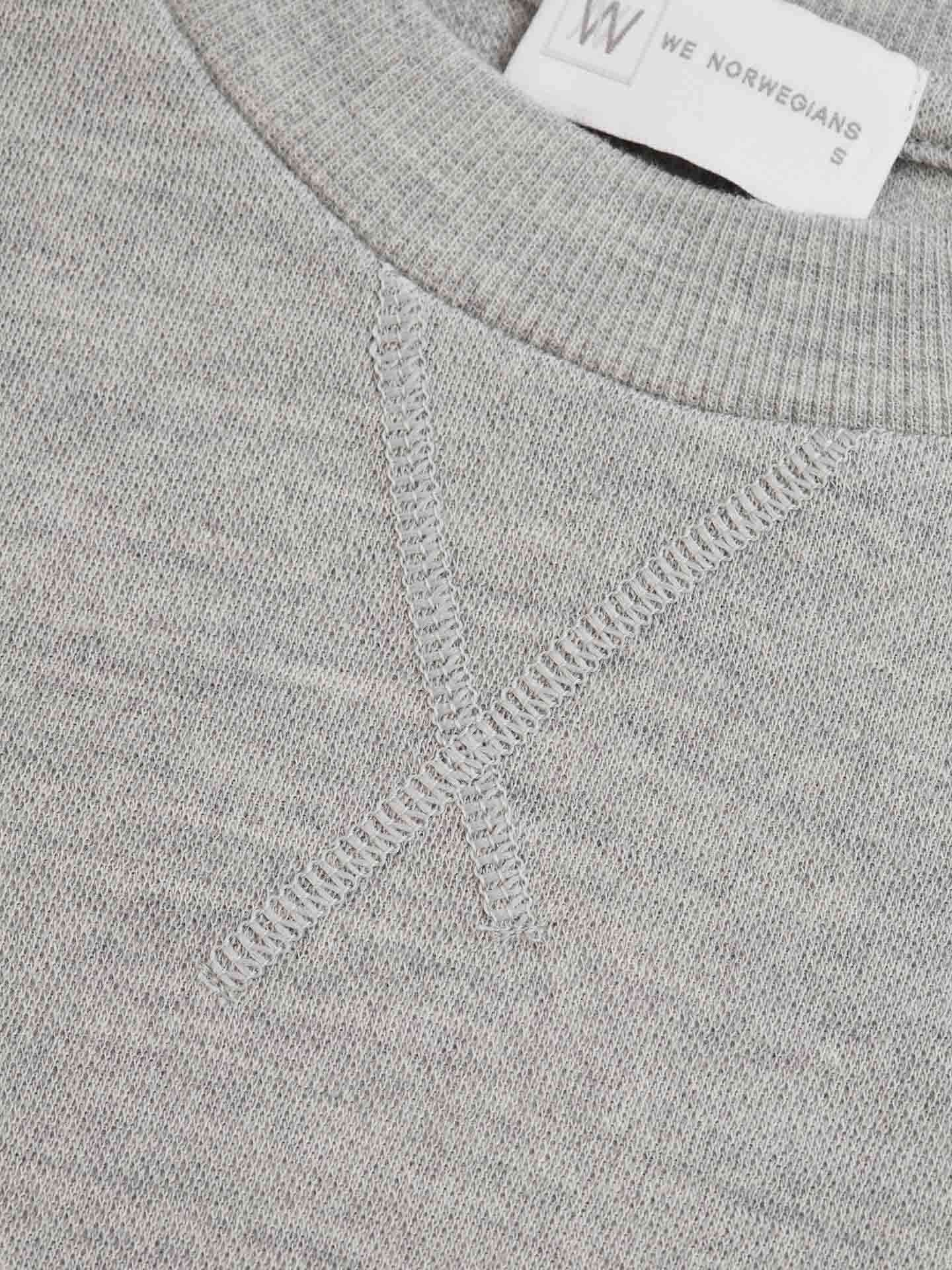 Close-up of the Tind Crewneck Women Grey Melange's neckline, showcasing detailed stitching forming an "X" pattern in a structured jacquard knit. The sweater's tag is partially visible at the top, displaying "We Norwegians" and the size "S." The fabric appears to be soft and finely woven Merino wool sportswear.