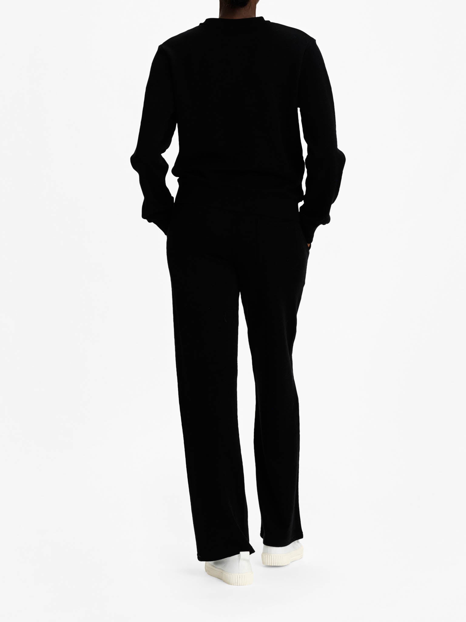 A person is standing with their back to the camera, dressed in an all-black sportswear outfit from We Norwegians, featuring the Tind Crewneck Women Black top and matching pants. They are also wearing white shoes. The background is plain white.