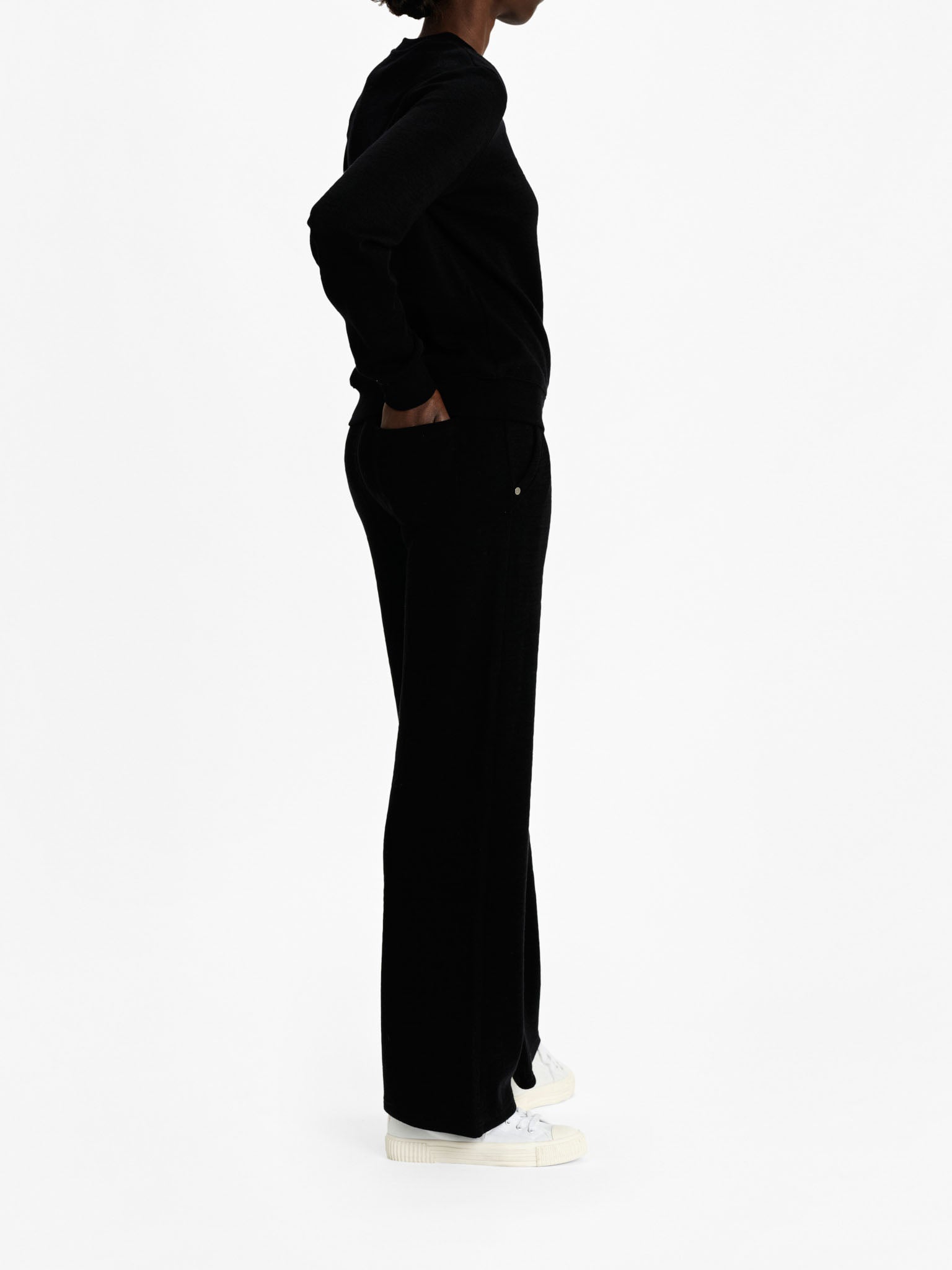 A person is standing in profile view against a plain white background. They are dressed in a black, long-sleeved Tind Crewneck by We Norwegians and black pants, with their hands in their pockets. They are also wearing white shoes.