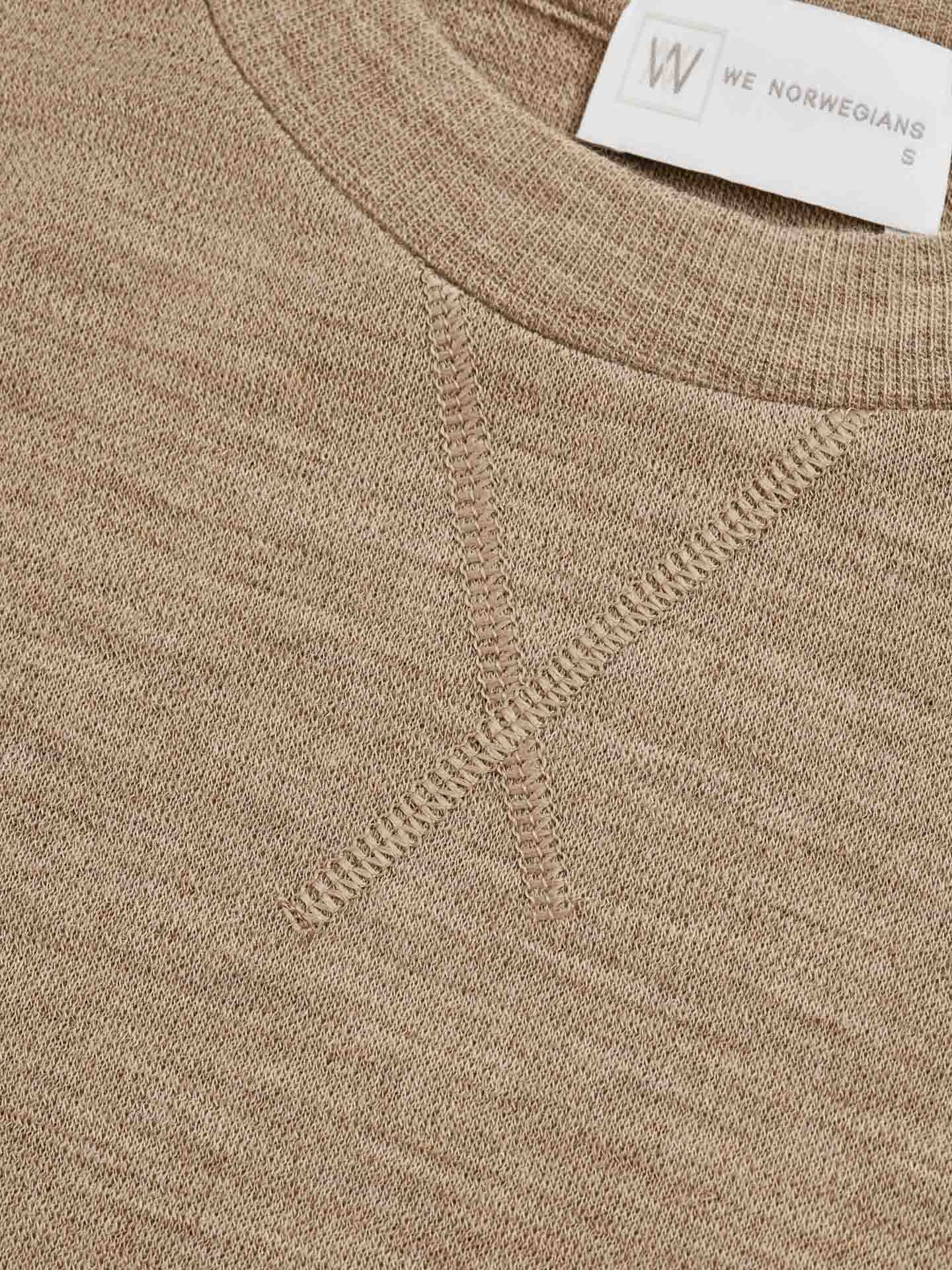 Close-up of a light brown knitted fabric made from pure Merino wool with visible stitching details and a white clothing label that reads "We Norwegians S". The stitching forms an "X" shape near the neckline, adding a subtle design element to the Tind Crewneck Women's Camel garment.