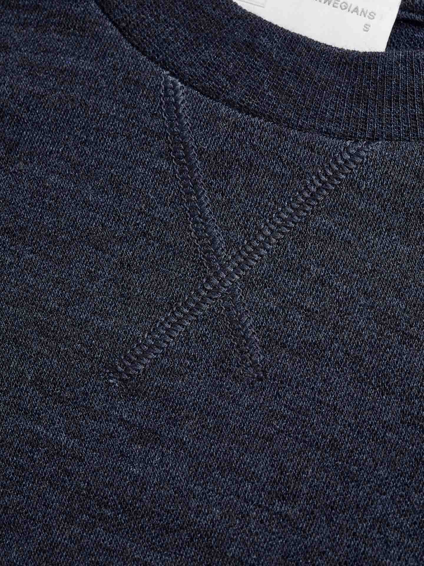 Close-up of the Tind Crewneck Women Navy by We Norwegians, a dark navy blue sweatshirt featuring a structured jacquard knit pattern. The luxurious sporty style is highlighted by the ribbed crew neckline and detailed "X" stitching near the collar. Part of a small white label with text is visible at the top right corner.