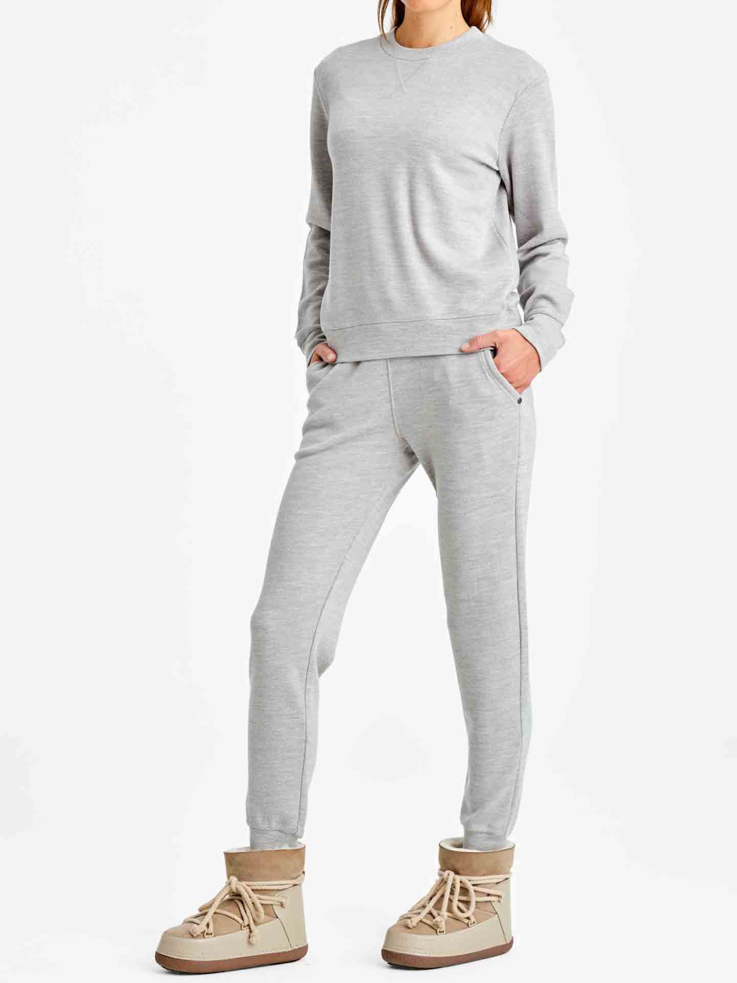 A person is standing against a white background wearing a matching light gray sweat suit from We Norwegians. The outfit includes a long-sleeved Tind Crewneck sweatshirt and Tind Jogger sweatpants from the Limited Time Bundle: Tind Crewneck & Tind Joggers Women. They are also wearing beige lace-up boots, with their hands in the pockets of the sweatpants.