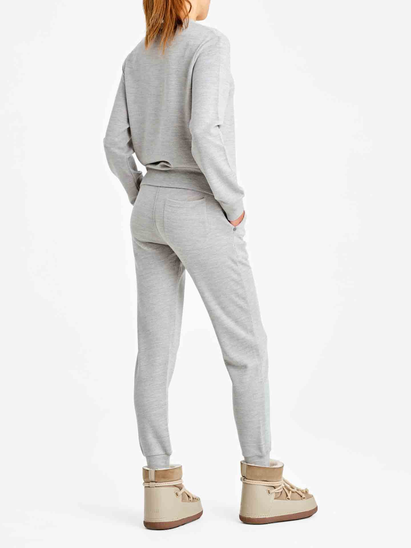 A person stands facing away, showcasing a luxurious sporty style with a casual outfit consisting of the Tind Crewneck Women Grey Melange sweatshirt by We Norwegians and matching joggers. The individual has one hand in a pocket and wears beige high-top boots, against a plain white background.