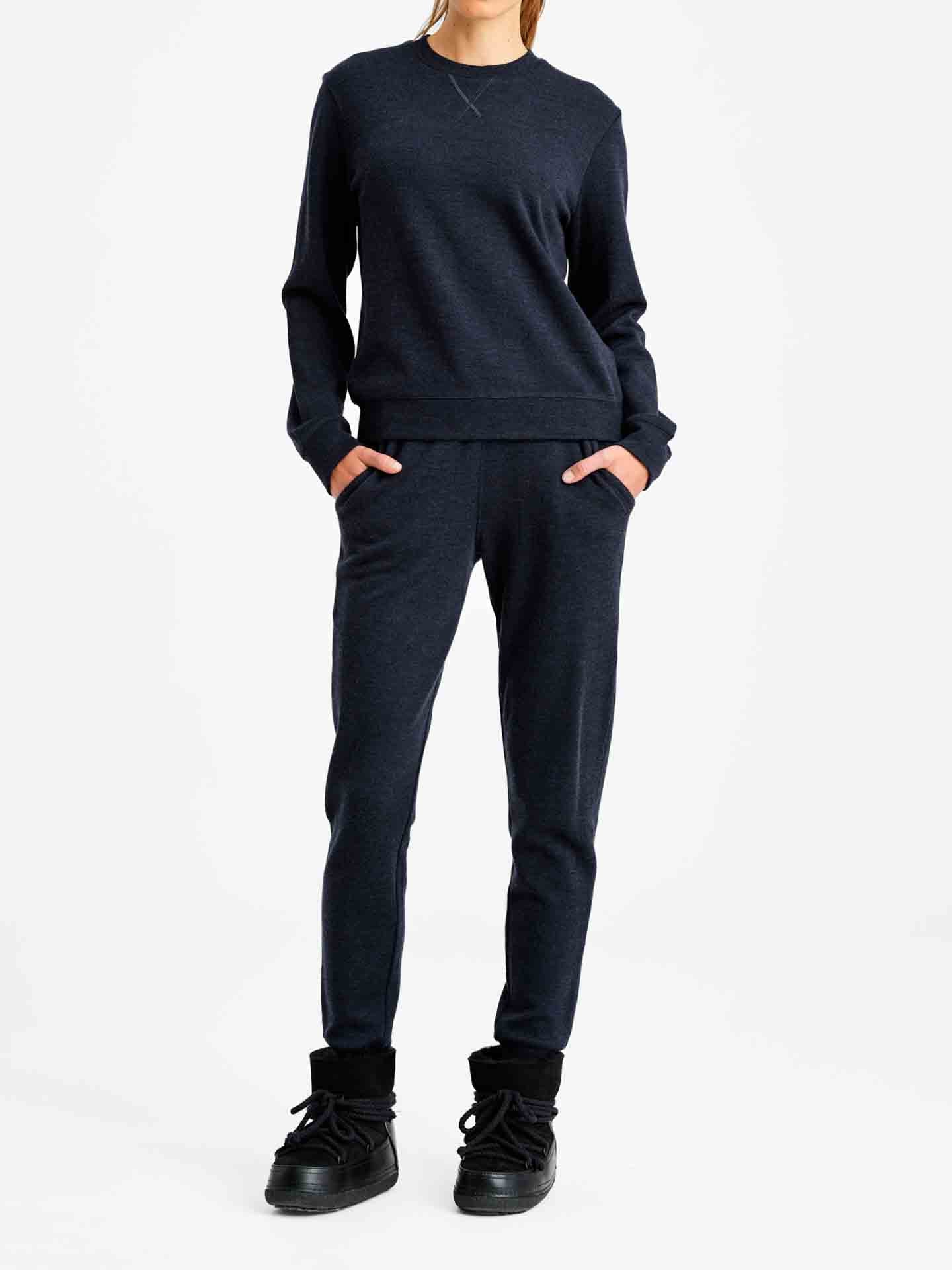 A person dressed in a matching dark-colored We Norwegians Limited Time Bundle: Tind Crewneck & Tind Joggers for women, with hands in pockets, standing against a plain white background. The individual is also wearing black ankle-high sneakers, giving the outfit a casual and comfortable look.