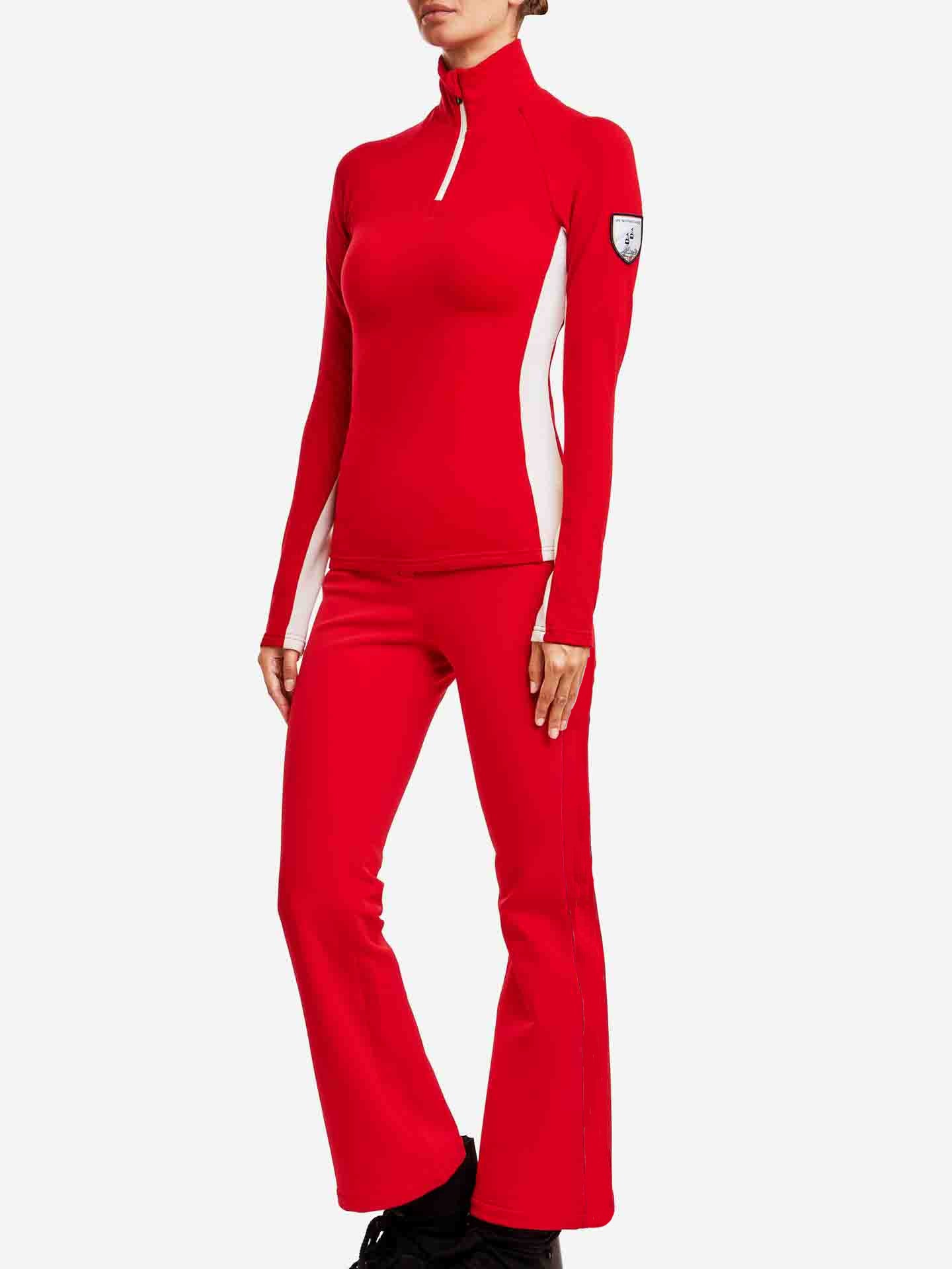 A person is wearing a red and white ski outfit, consisting of a long-sleeve top with a half-zip collar and matching We Norwegians Kvitfjell Bootleg Ski Pant for women. Made from brushed merino fleece with 4-way stretch soft shell, the outfit features white accents on the sides and a logo patch on the upper left arm. The person is standing against a white background.