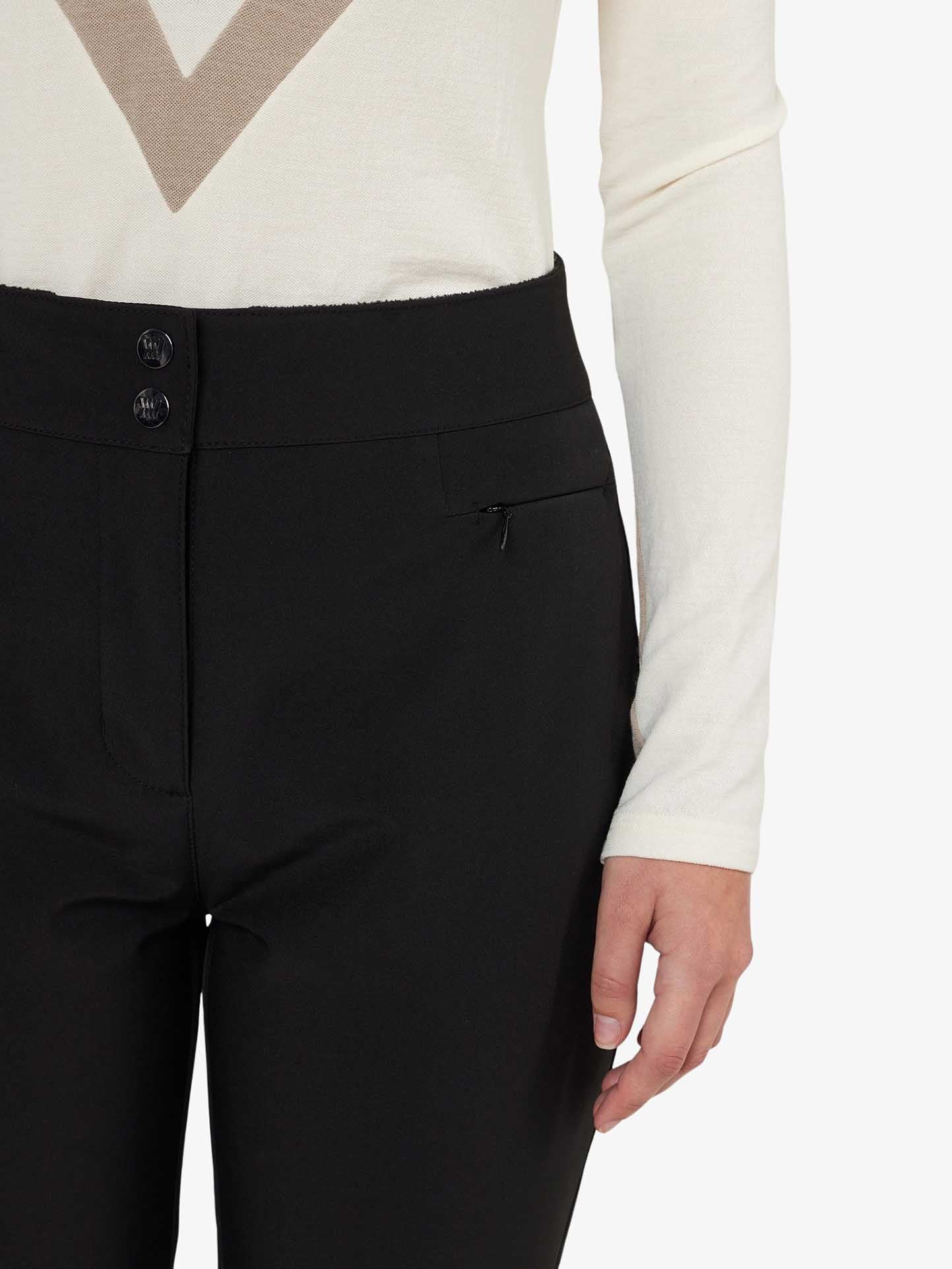 A person is wearing We Norwegians Kvitfjell SlimLeg SkiPant Women in sleek black, high-waisted with stirrups and two buttons, paired with a cream long-sleeve top featuring a geometric neckline design. The pants also have a small zipper pocket.