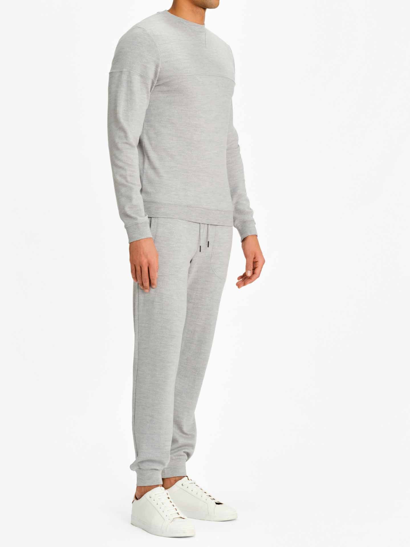 A person is wearing a light gray, long-sleeved Tind Crewneck Men sweatshirt by We Norwegians, paired with matching light gray Tind Joggers and white sneakers. The outfit is comfortable and casual. The background is plain white. The person is standing and partially visible from a side view.