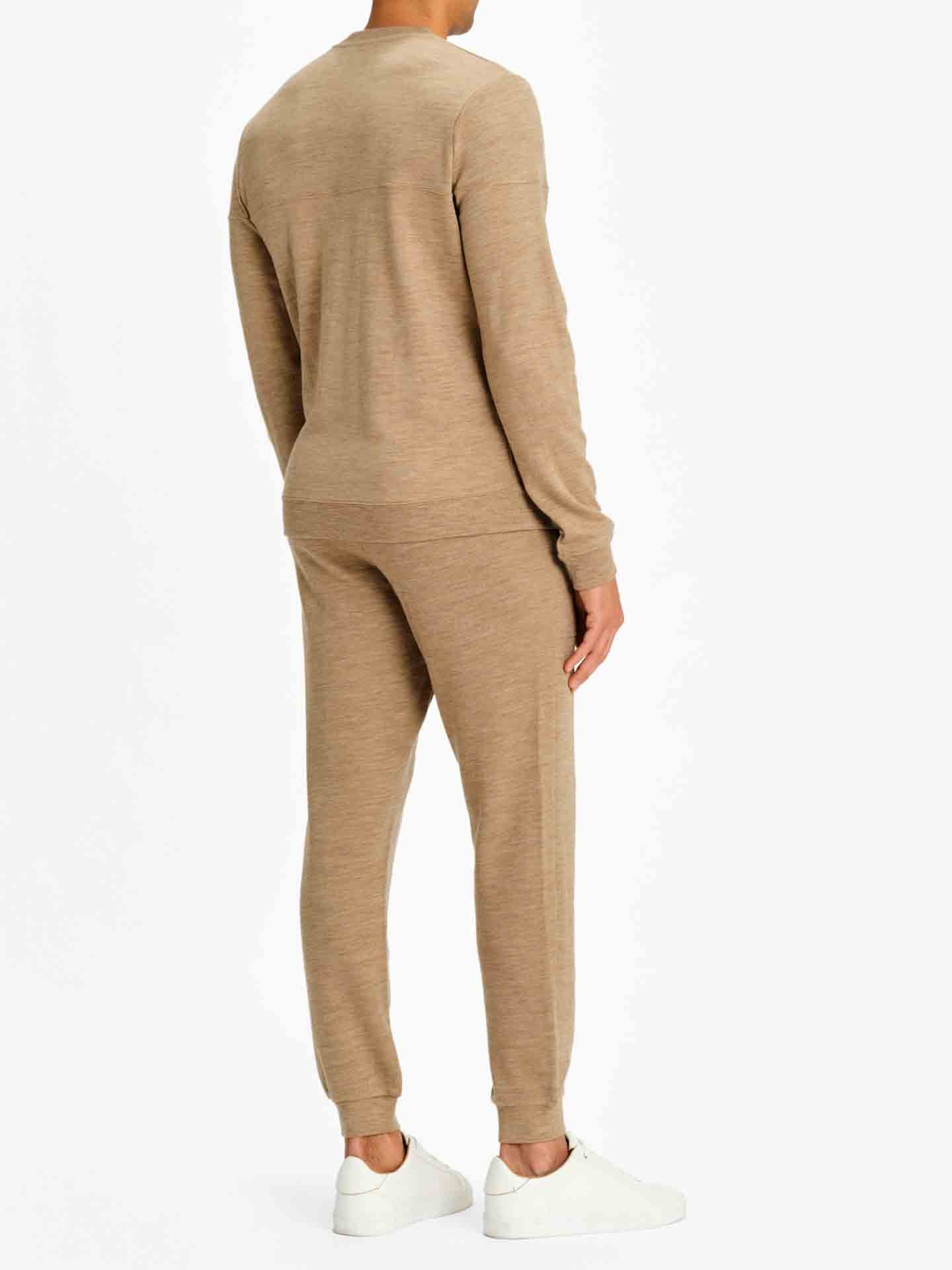 A person stands against a plain white background, facing away. They are wearing a tan long-sleeve top called the Tind Crewneck Men Camel by We Norwegians and matching Tind Joggers made of Merino wool, along with white sneakers. The sportswear outfit appears comfortable and casual.