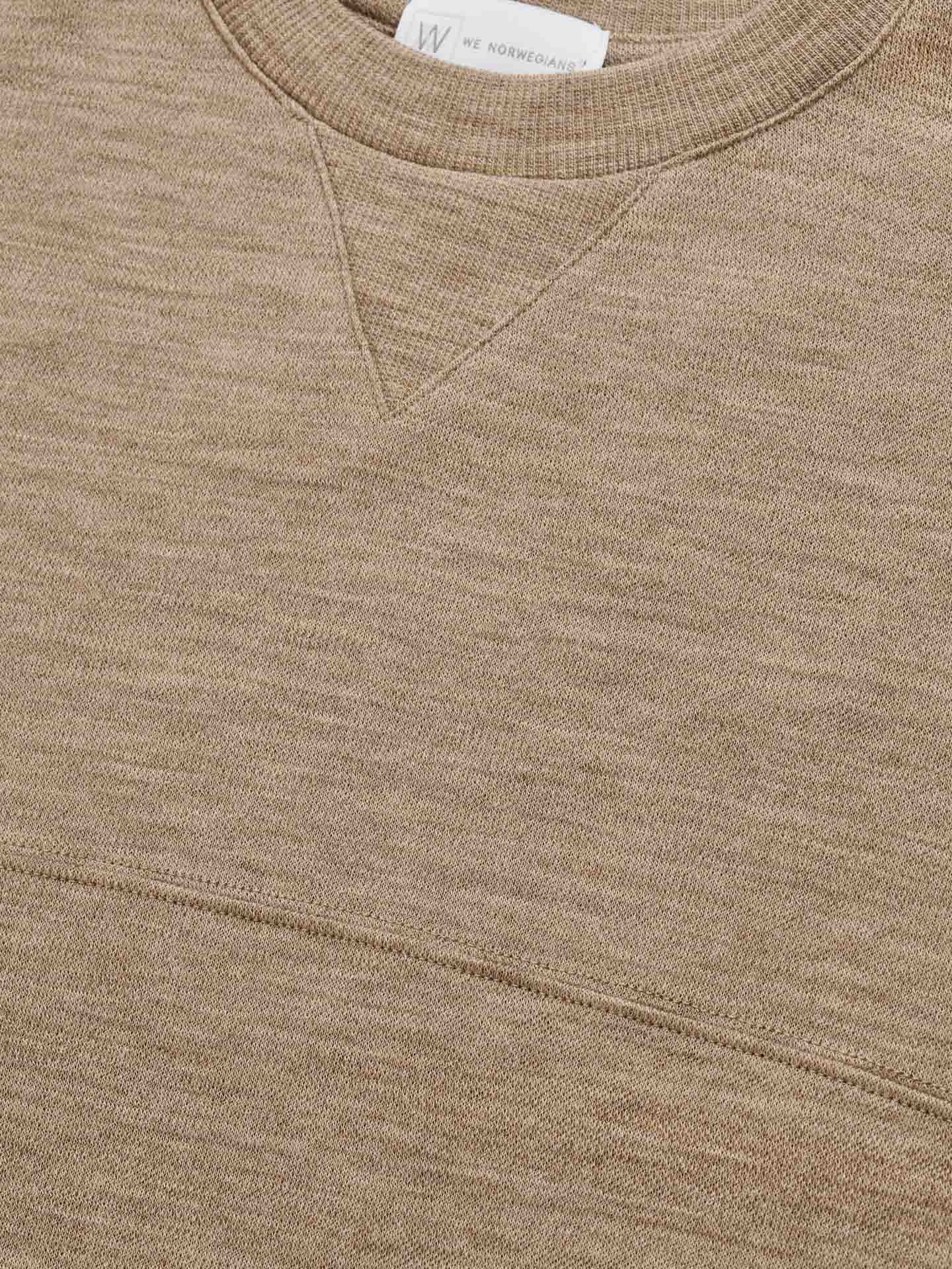 Close-up of the Tind Crewneck Men Camel from We Norwegians, showcasing a beige knit design with a ribbed crew neckline and a small 'V' detail below the collar. A white tag with text is partially visible inside the collar. Crafted from soft Merino wool, this sweater offers a textured appearance similar to that of Tind Joggers in both comfort and style.