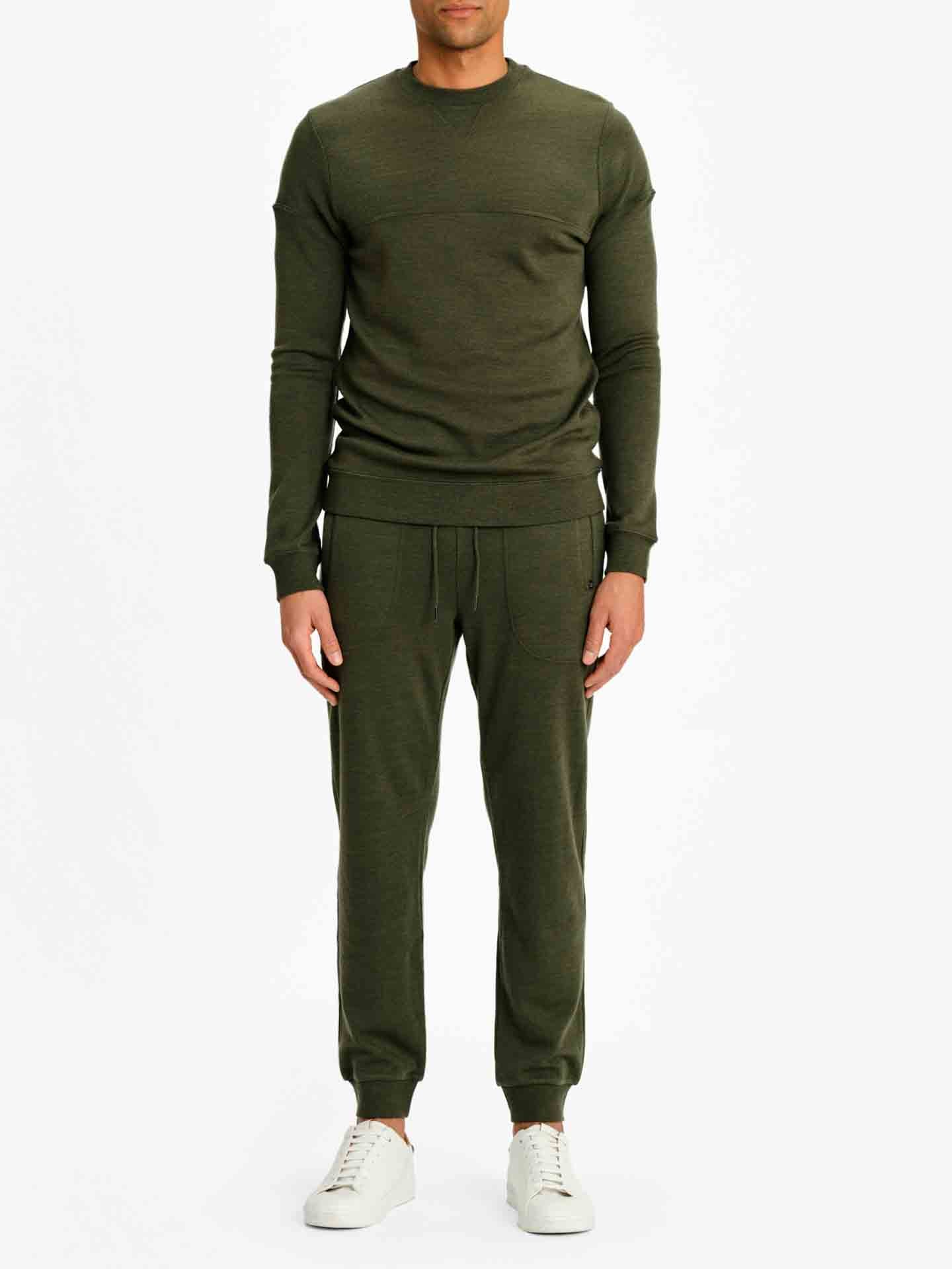 A man stands against a plain white background wearing the We Norwegians Tind Crewneck Men Olive Green jacquard knit long-sleeve sweater and jogger pants set, paired with white sneakers. The outfit appears comfortable and casual. The man's face is not visible in the image.