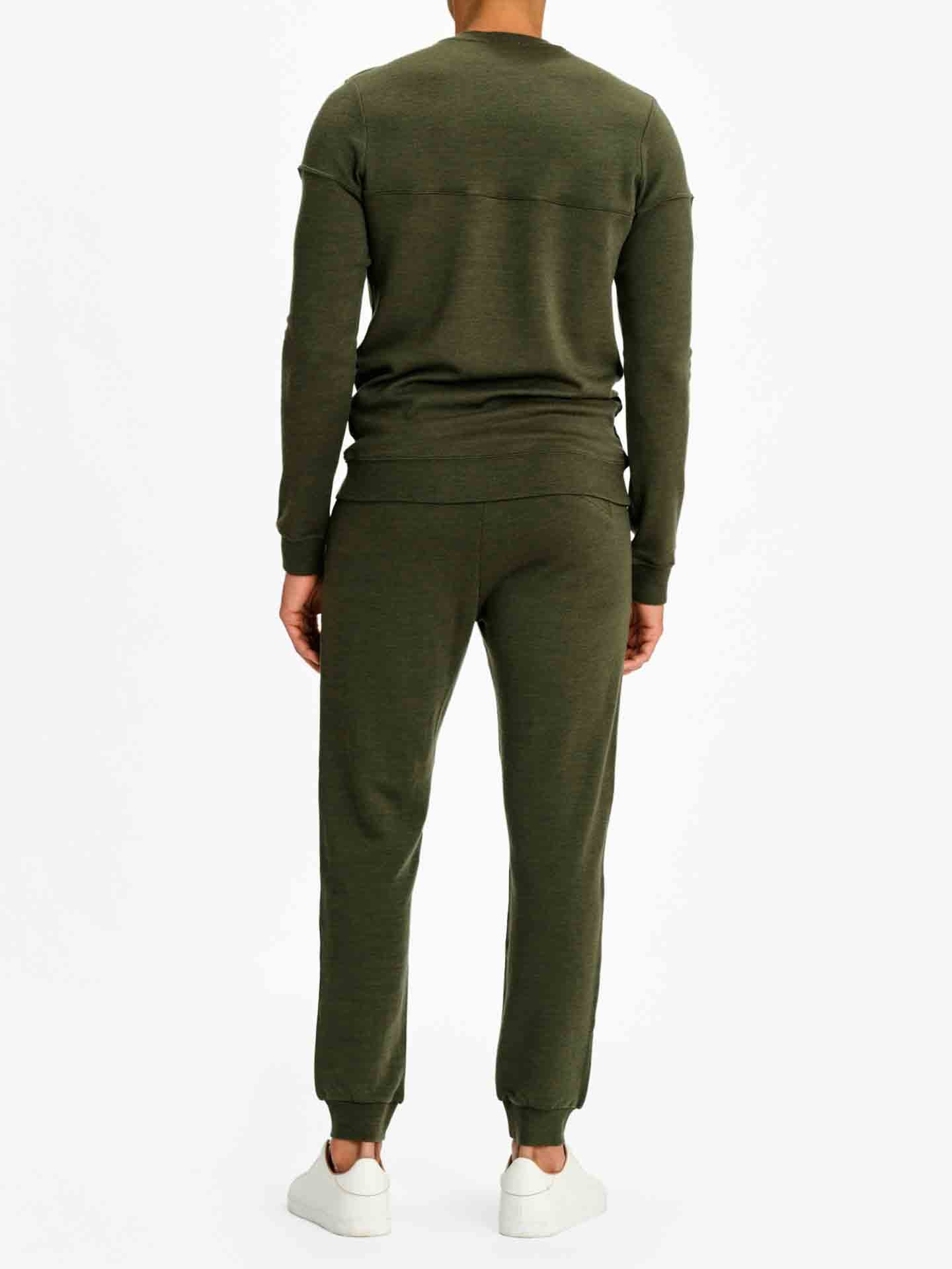 A person is standing with their back to the camera, dressed in a matching We Norwegians Tind Crewneck Men Olive Green sweatsuit that exudes classic sportswear vibes, featuring a long-sleeve top and jogger pants. They are also wearing white sneakers. The background is plain white.